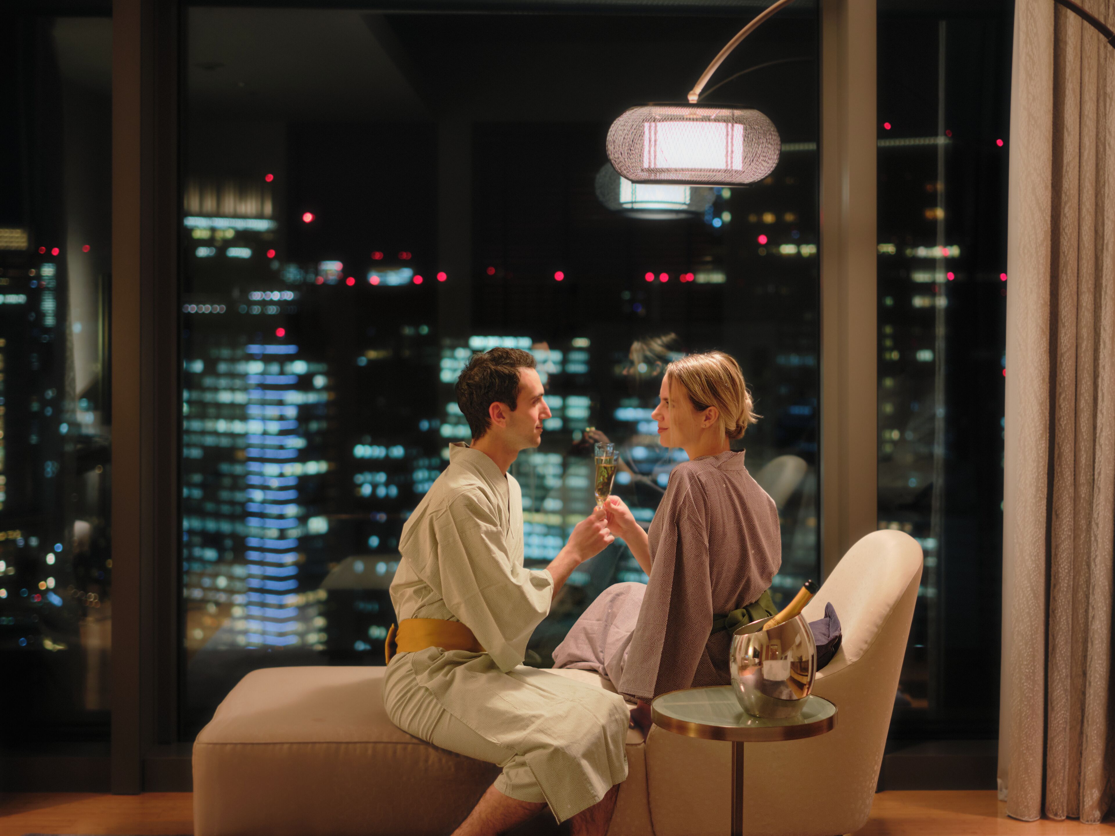 Una pareja sentada en un sofá frente a una vista panorámica de la ciudad, con una mesa entre ellos que sostiene una botella de champán y copas.