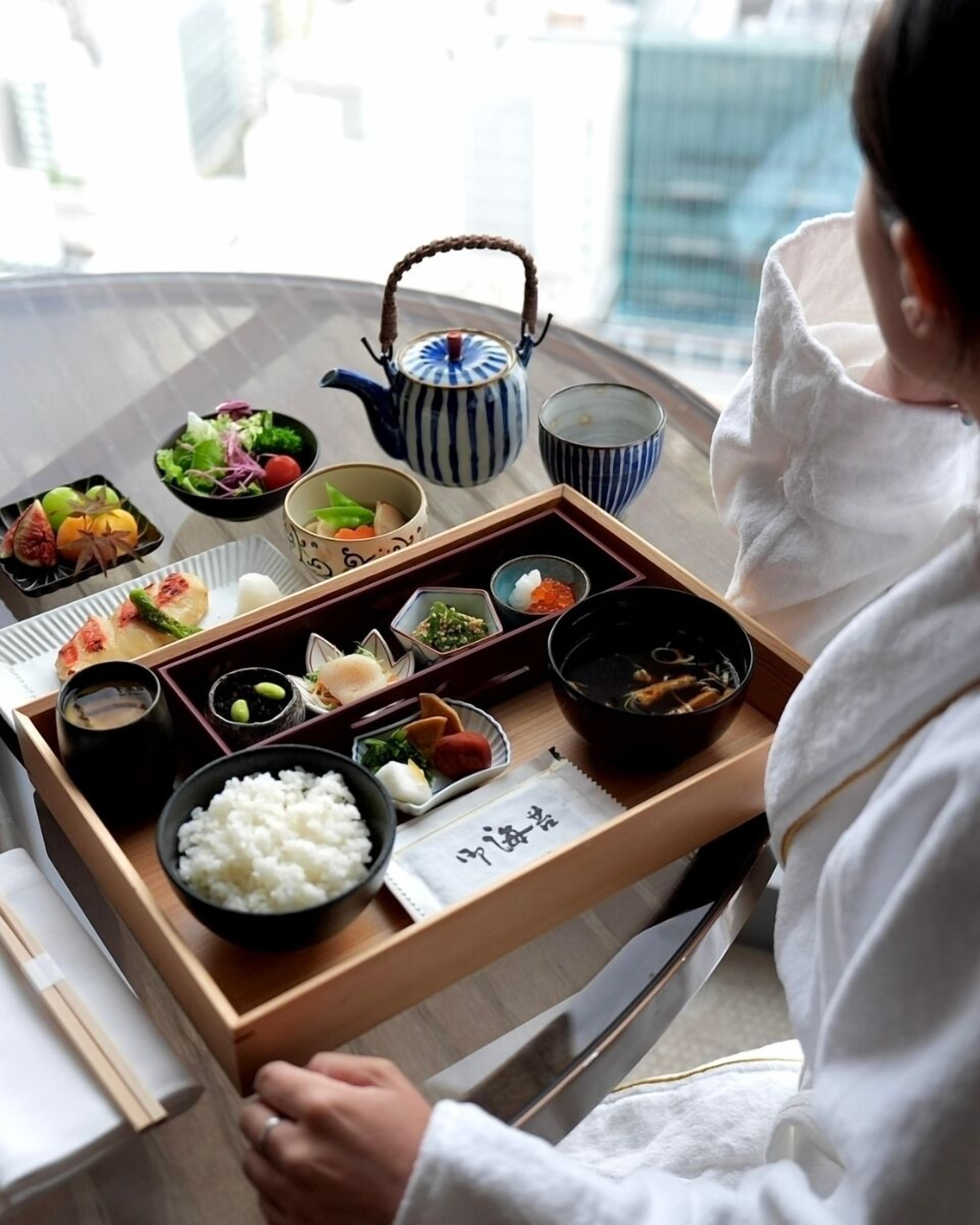 A tray with various Japanese dishes, including rice, vegetables, and fish, on a table with a teapot and cups.