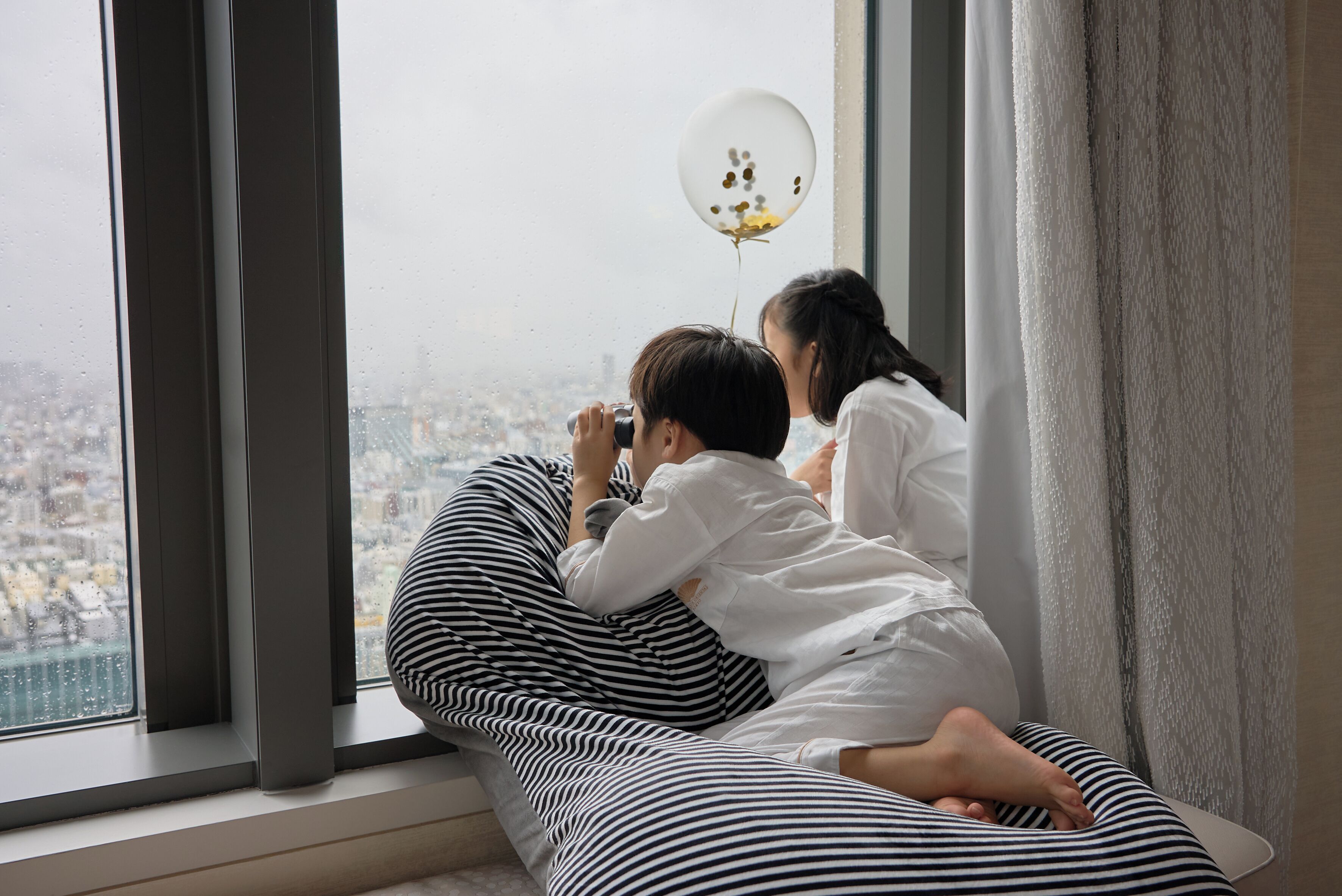 Two individuals sitting on a striped bed by a window with a view of a cityscape.