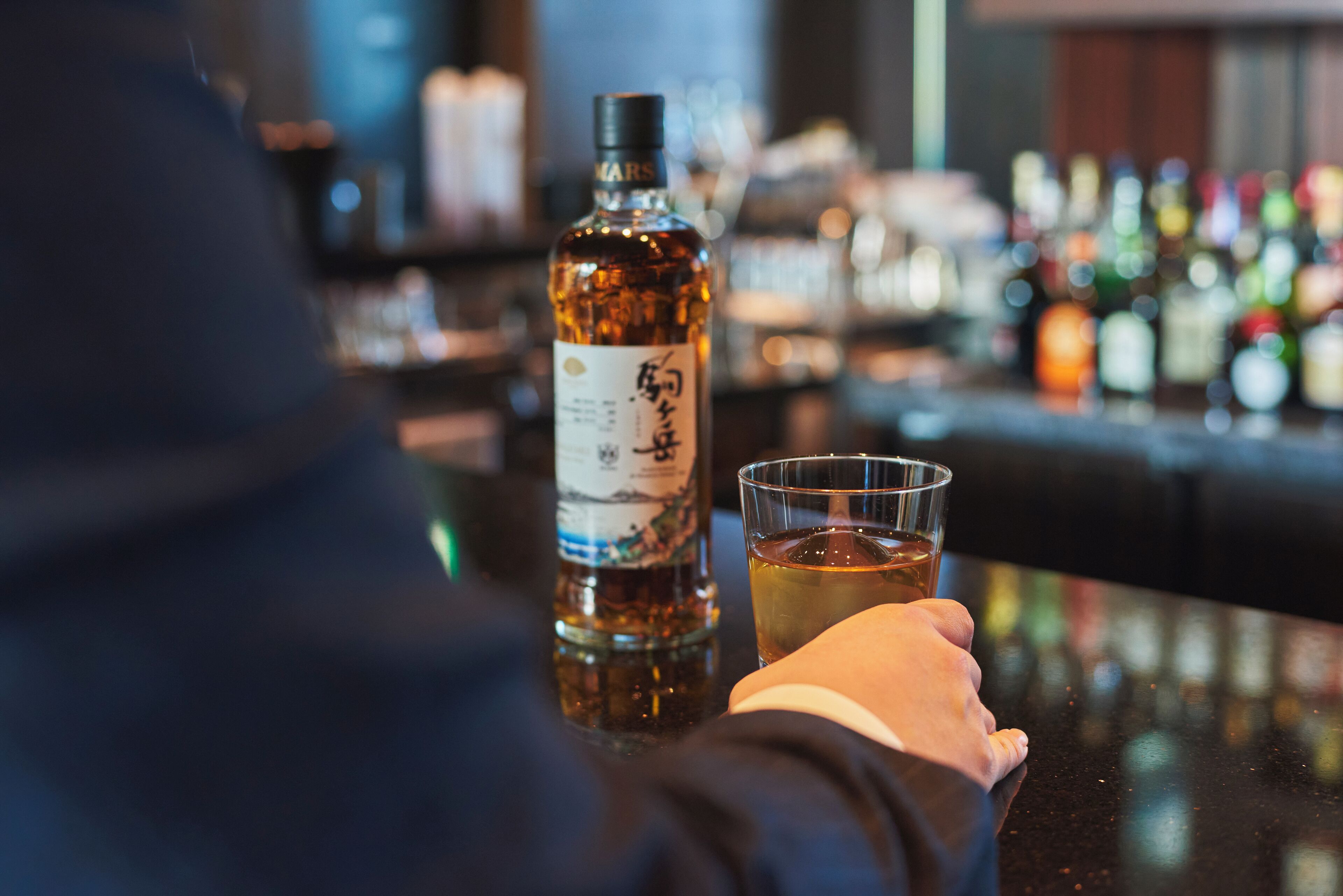 Una persona sosteniendo una copa de whiskey y una botella con etiqueta, en un bar con varias botellas al fondo.