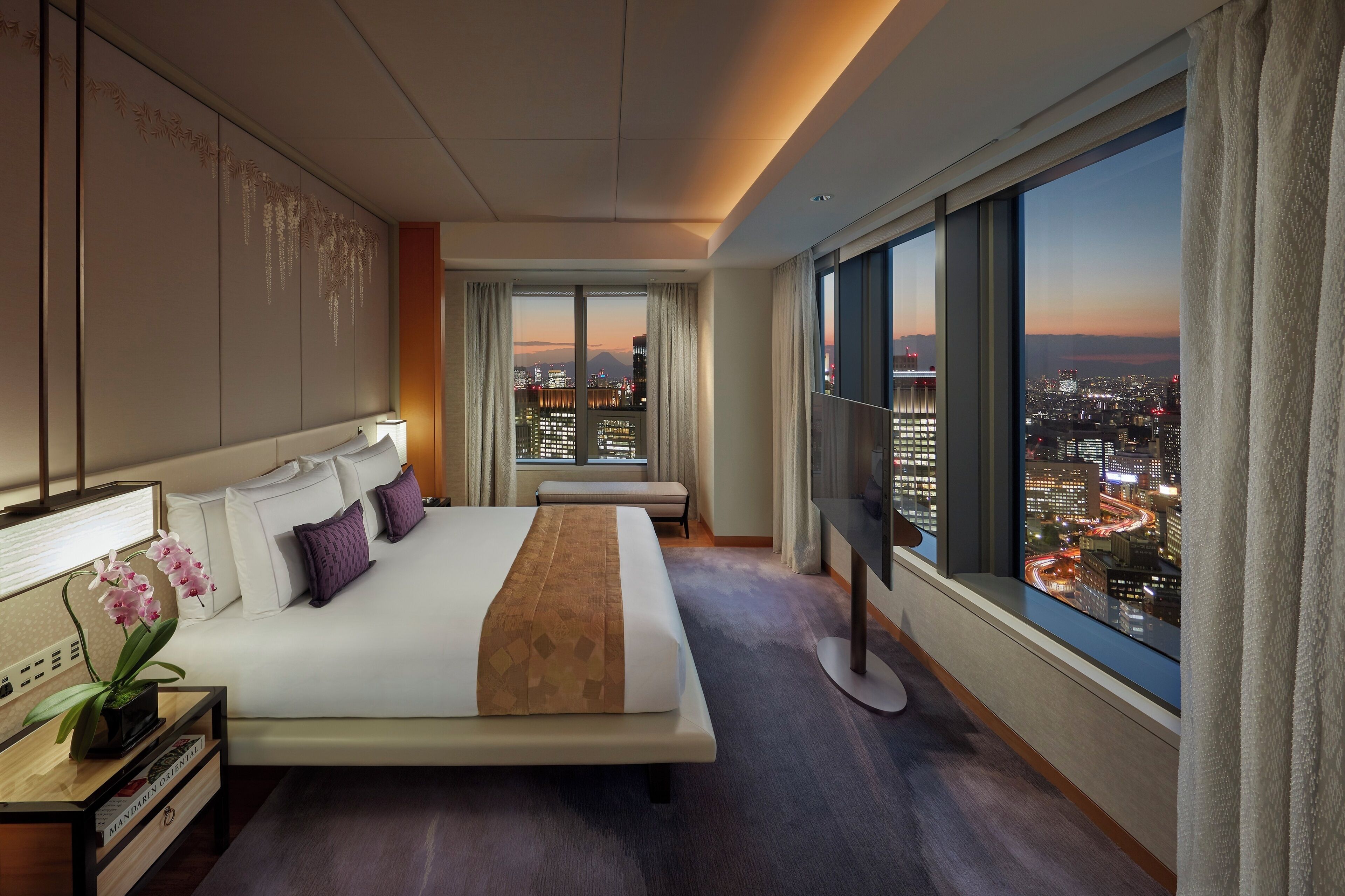 A hotel room with a large bed, a TV, a desk with a lamp, a window with a city view, and a wall with a decorative panel.