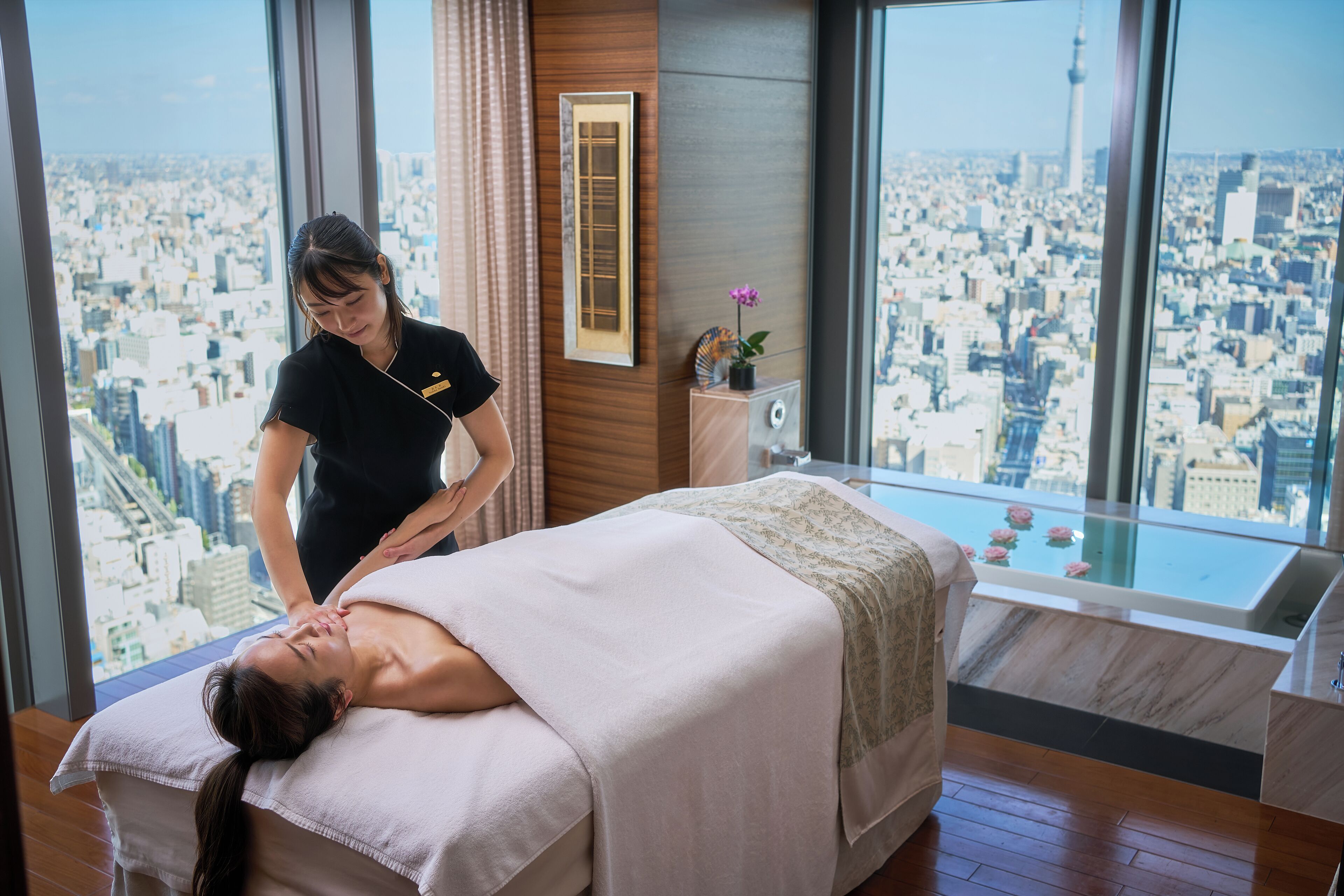 A massage therapy room with a city view, a massage table, a small side table with a plant, and a large window.