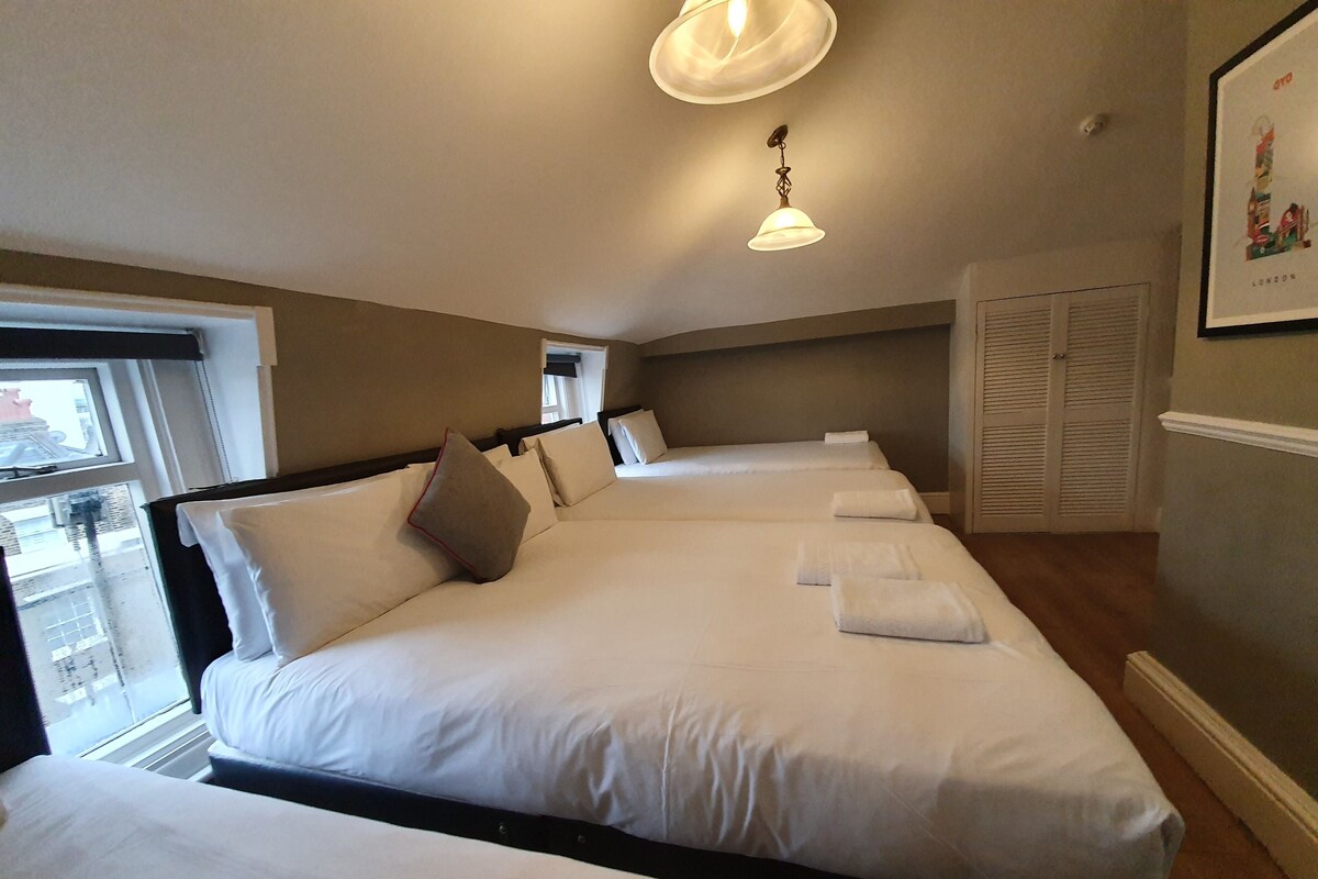 A hotel room with three beds, a window with blinds, a ceiling light, and a framed picture on the wall.