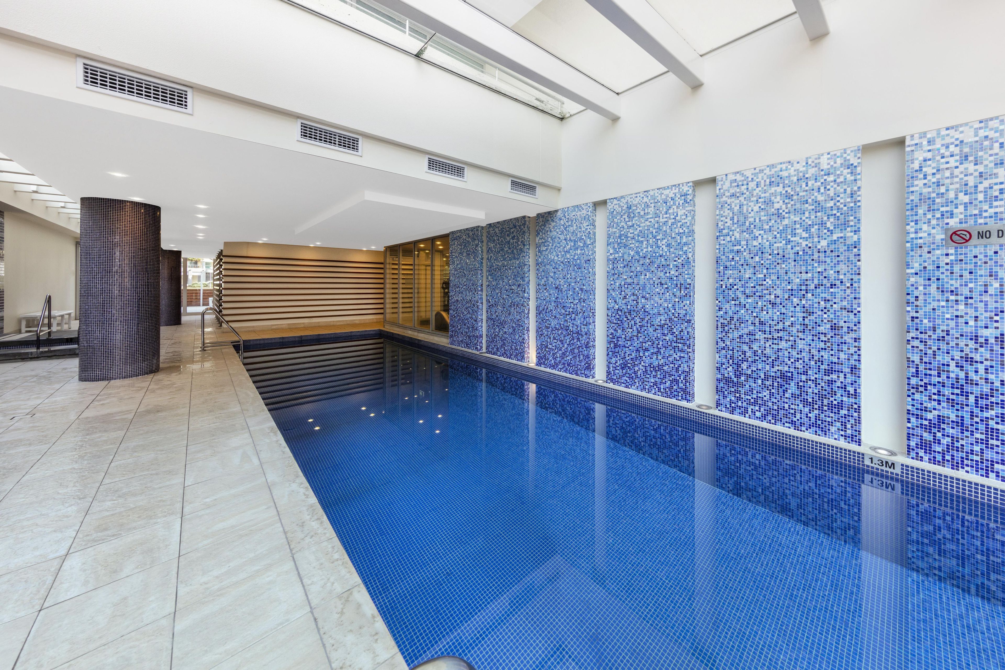Ein Hallenbad mit blau gefliestem Boden und Wänden, einem Sprungverbotsschild und einer Wand mit blauem Mosaikmuster.