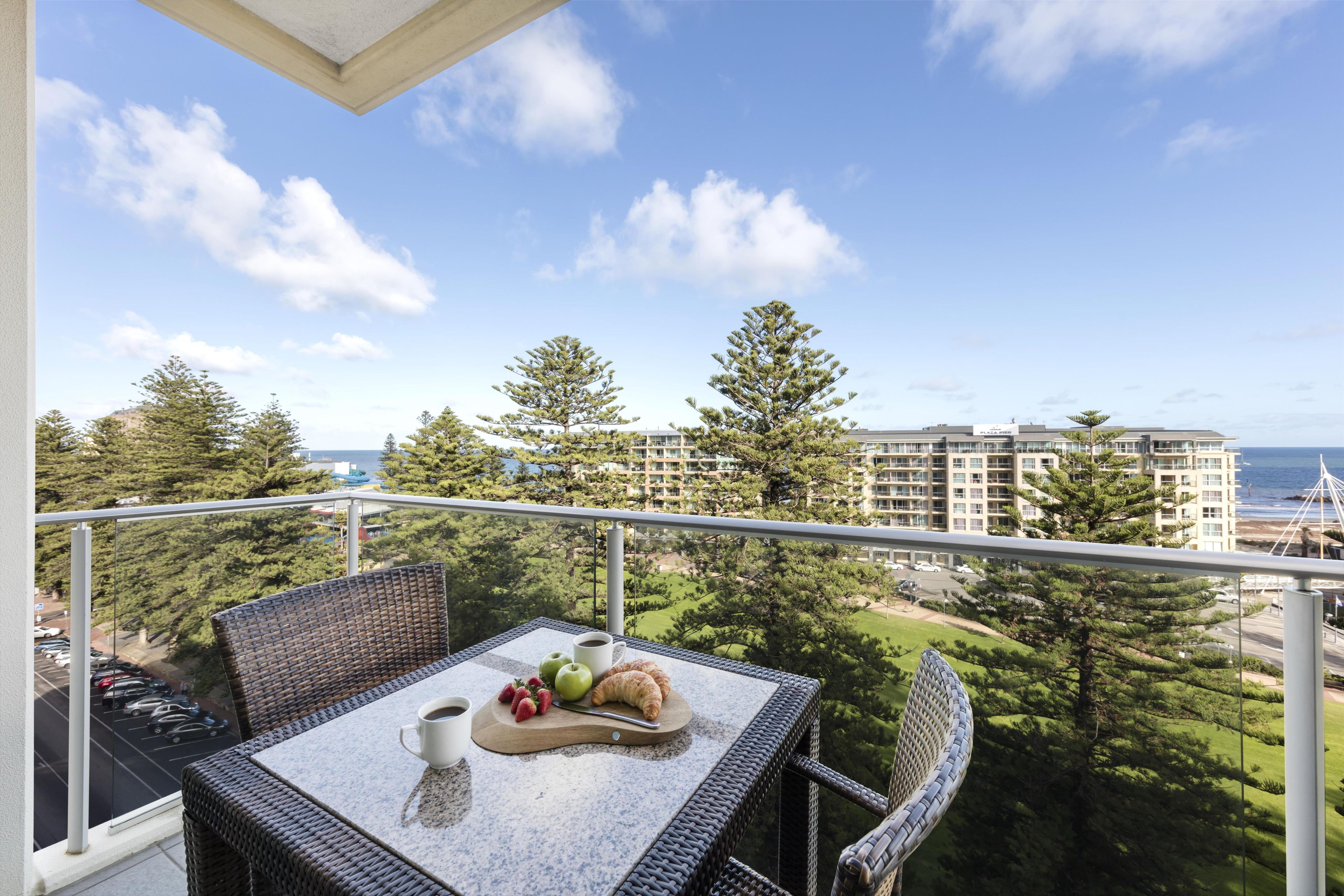 Un balcon avec des meubles en osier, une table garnie de fruits, une tasse de café et une vue sur un bâtiment situé sur la côte.
