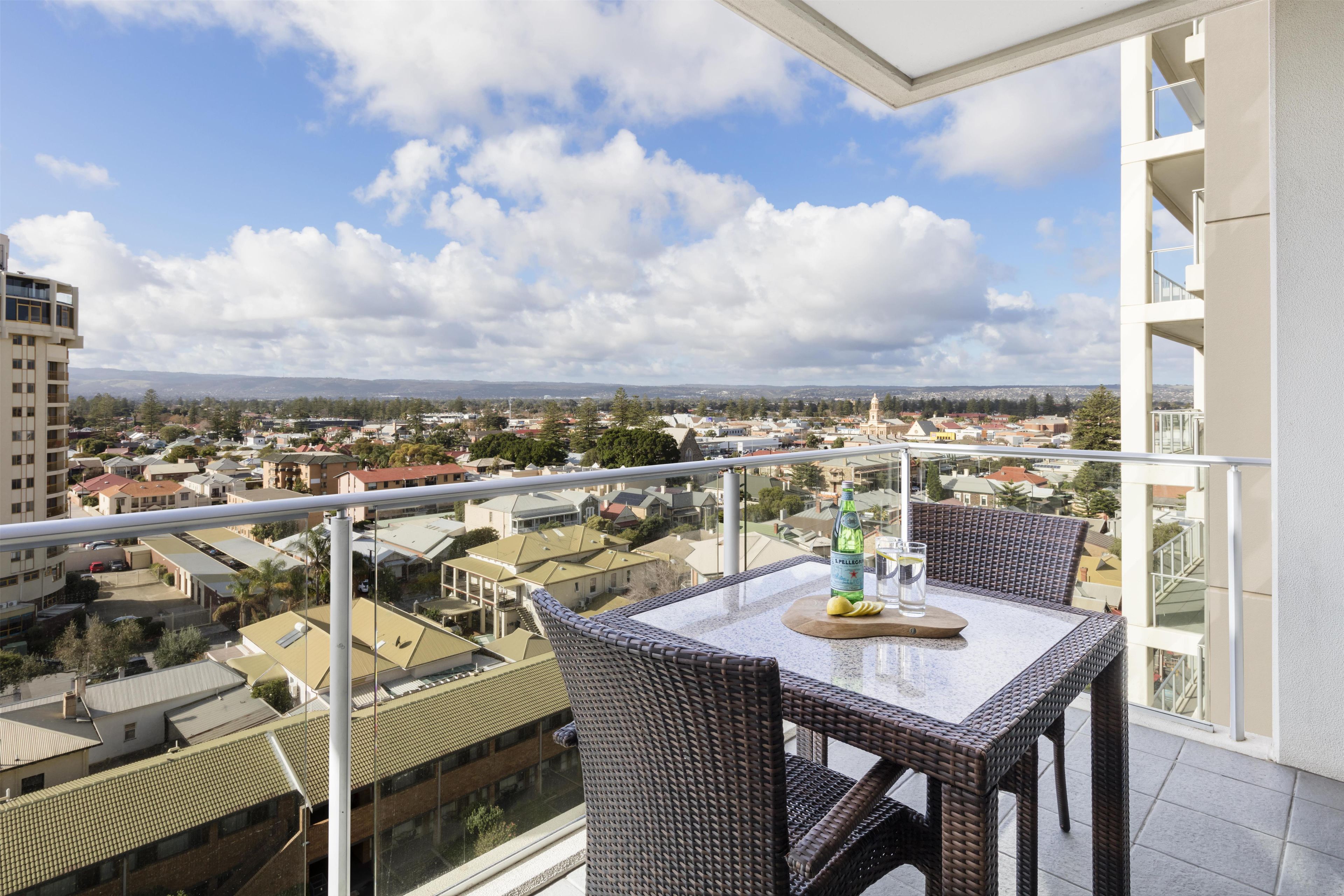 A balcony with a table and chairs, overlooking a residential area with various buildings and a clear sky.