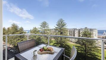 A balcony with wicker furniture, a table with a croissant, strawberries, and a cup of coffee, overlooking a coastal view with buildings and trees.