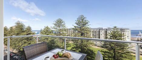 Un balcon agrémenté de meubles en osier, une table avec un croissant, des fraises et une tasse de café, offrant une vue sur la côte, avec ses bâtiments et ses arbres.