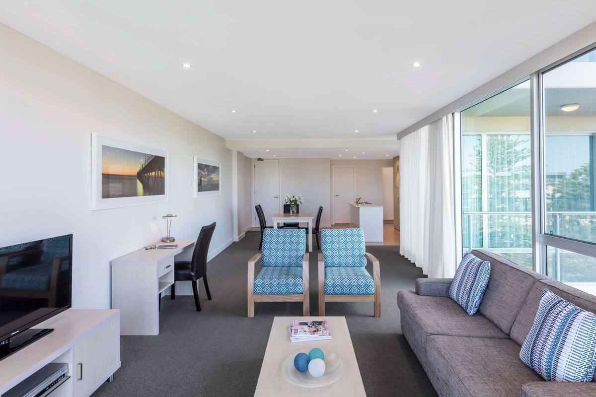 Une chambre d’hôtel moderne dotée d’une télévision à écran plat, d’un canapé, de deux fauteuils à motifs, d’un coin repas avec une table et des chaises, et de grandes fenêtres offrant une vue sur la ville.