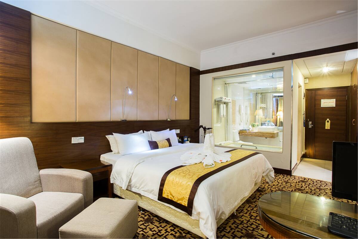 A hotel room with a large bed, a sitting area with a chair, a desk with a computer, and a bathroom visible through a glass door.