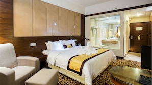 Habitación de hotel con una cama grande, una zona de estar con una silla, un escritorio con ordenador y un baño visible a través de una puerta de cristal.