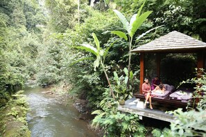 A wooden gazebo with a thatched roof overlooking a river, surrounded by lush greenery. - Hanging Gardens of Bali (Payangan)