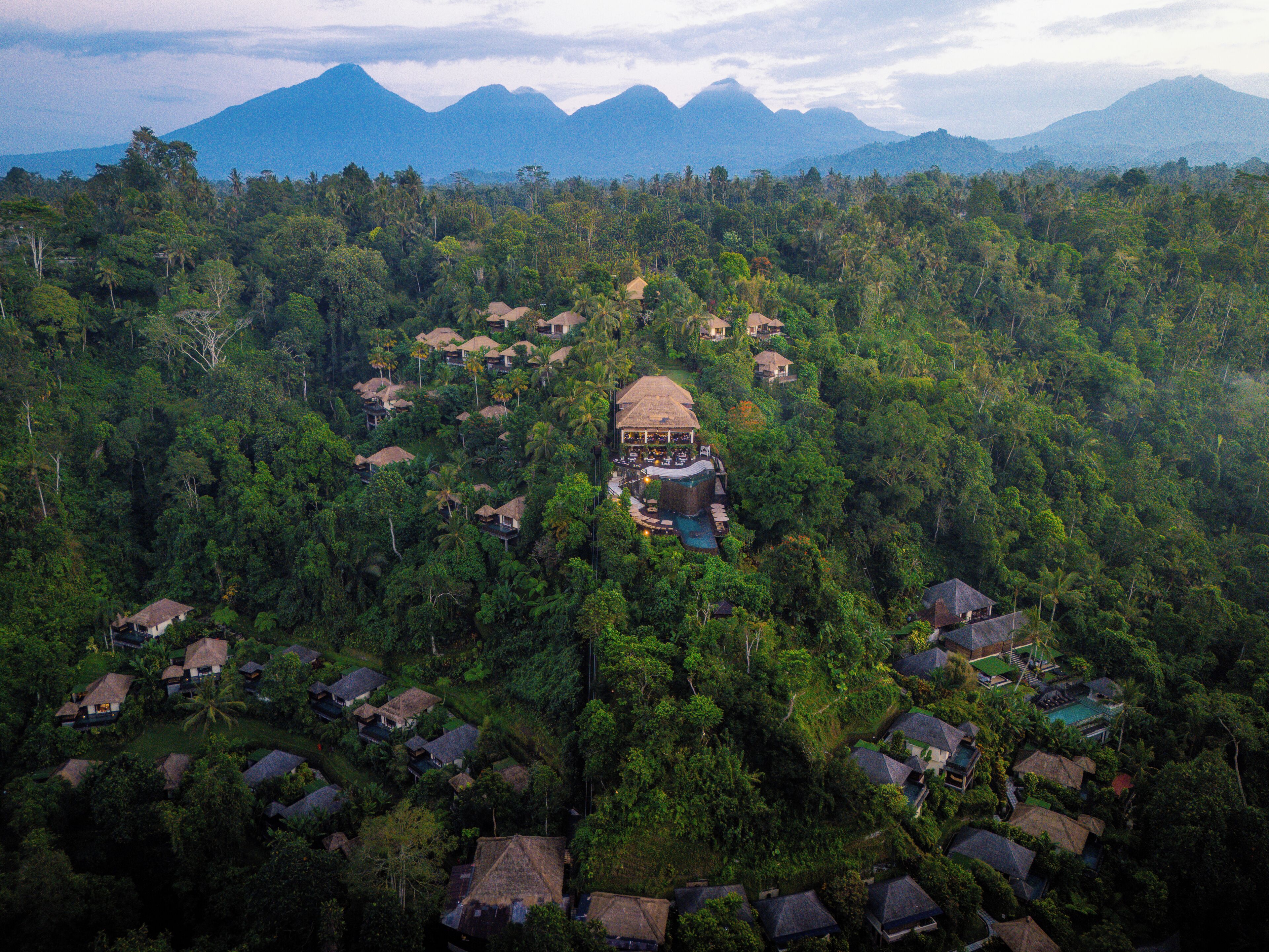 An aerial view of a resort with multiple bungalow-style accommodations surrounded by dense forest, a central building with a pool, and mountains in the background.
