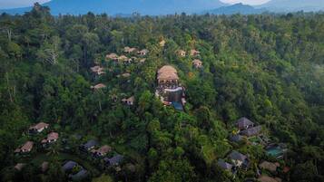 An aerial view of a resort with multiple bungalow-style accommodations surrounded by dense forest, a central building with a pool, and mountains in the background.