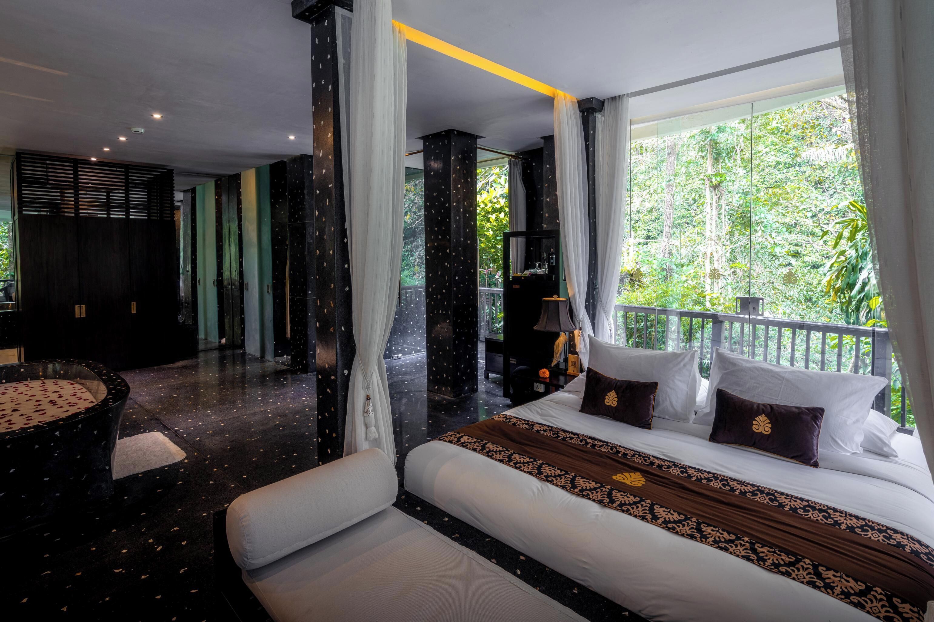 A hotel room with a balcony, a bed with decorative pillows, a seating area with a round table, and a view of greenery outside.