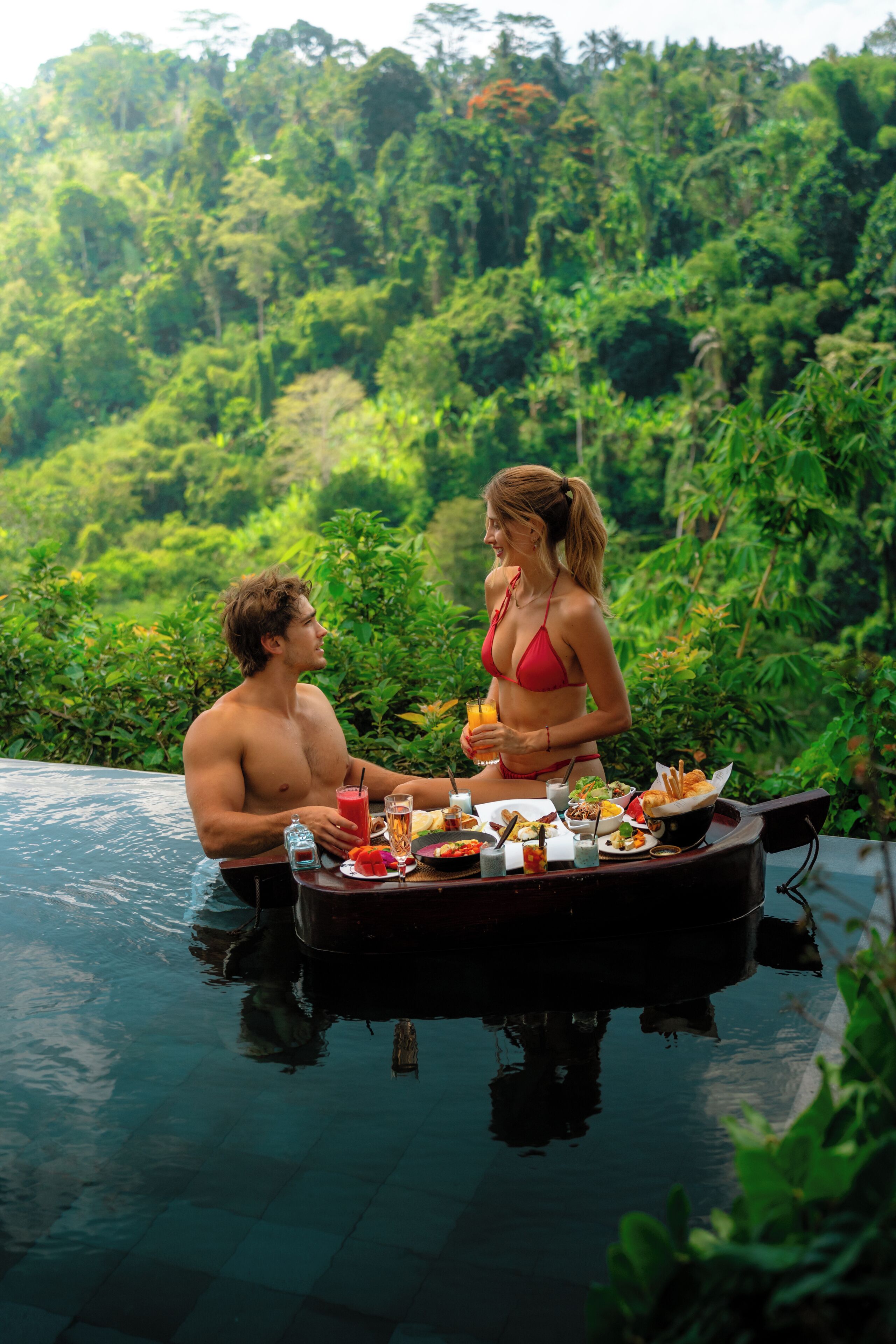 Una pareja cenando junto a una piscina con exuberante follaje de fondo.