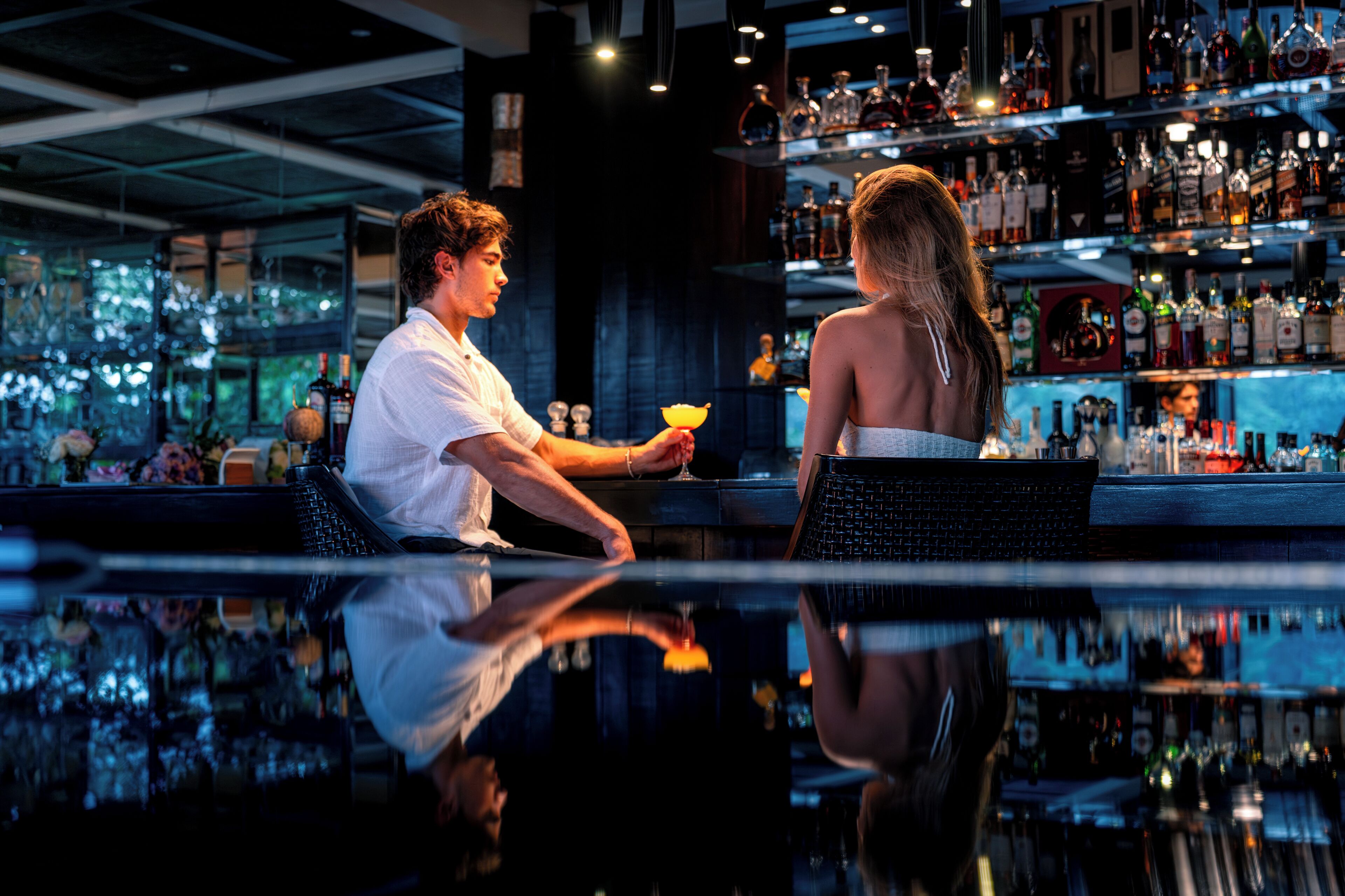 A bar with a polished floor reflecting the interior, shelves stocked with various bottles, and a person seated at the bar.