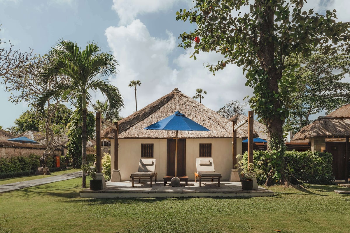 A thatched-roof building with two lounge chairs and a table in front, surrounded by greenery and palm trees.