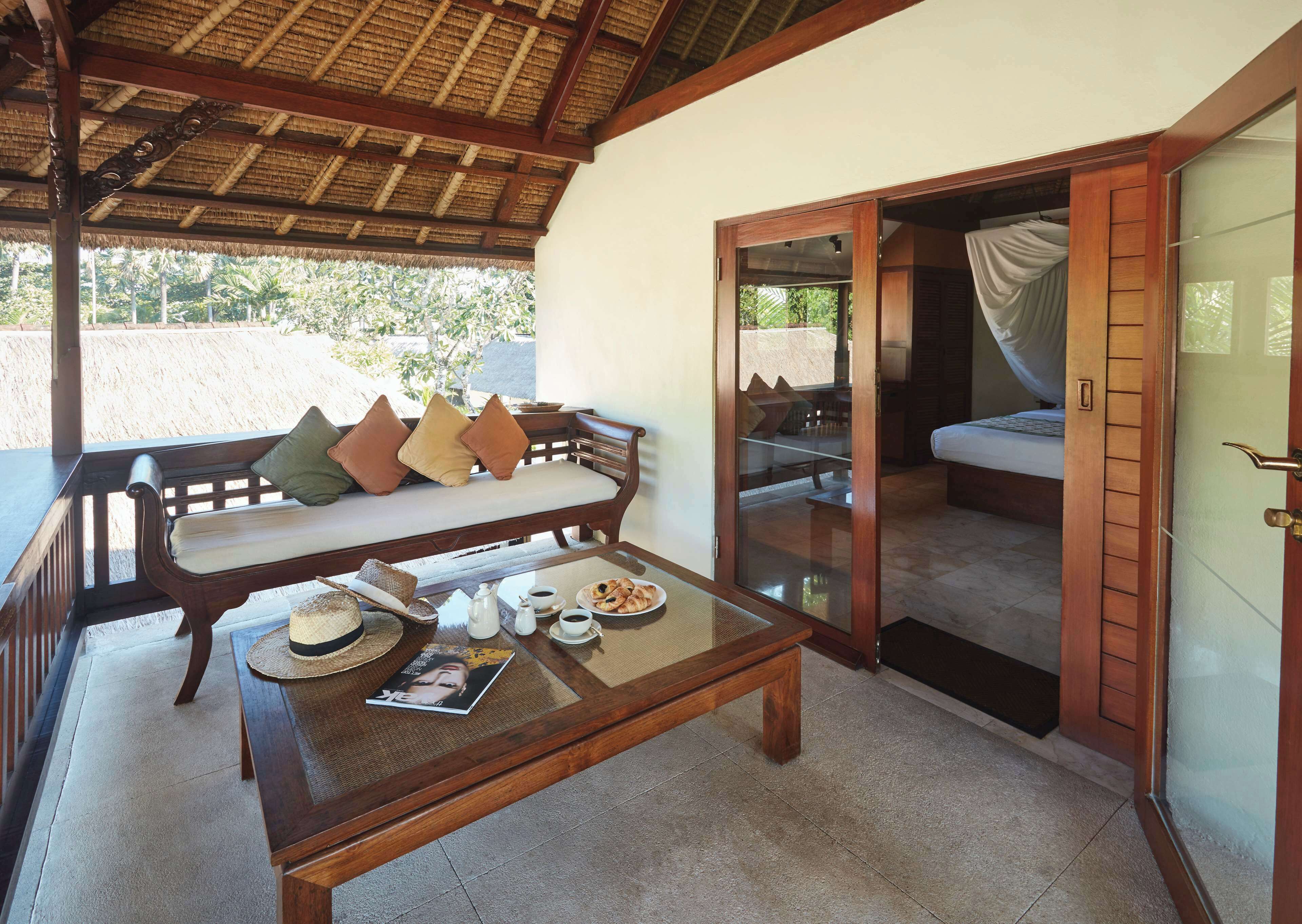 A wooden bench with cushions, a table with a magazine and a basket, and a bed with a canopy in a room with a thatched roof and sliding glass doors.