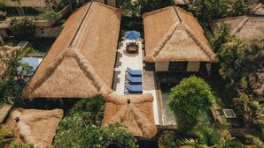 Vista aérea de un resort con edificios de techo de paja, una piscina central y abundante vegetación.