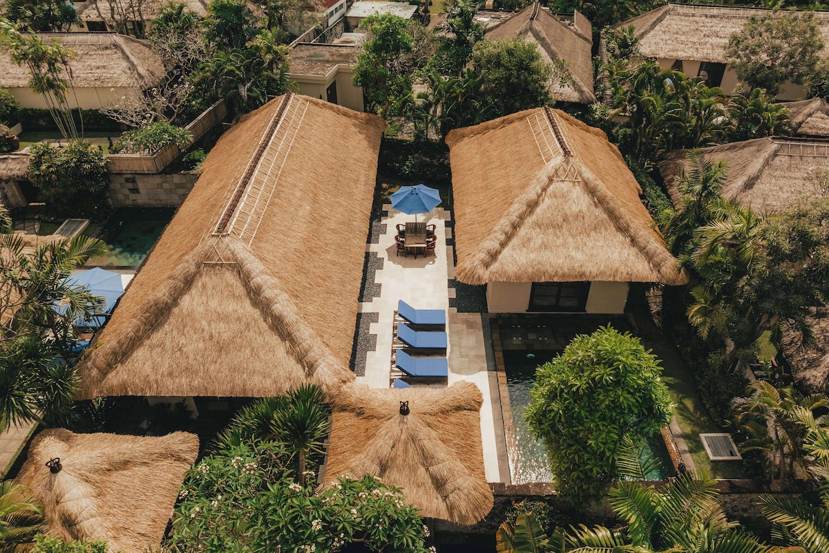An aerial view of a resort with thatched-roof buildings, a central pool, and lush greenery.