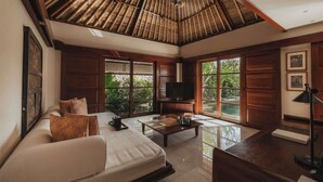 Amplia sala de estar con un sofá grande, una mesa de centro y una televisión. Tiene techo de madera y ventanas amplias que ofrecen vistas al verde exterior.