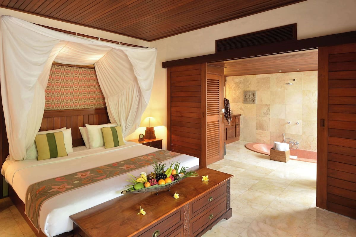 A bedroom with a bed, a bedside table, a fruit basket, and a bathroom with a sink and shower.