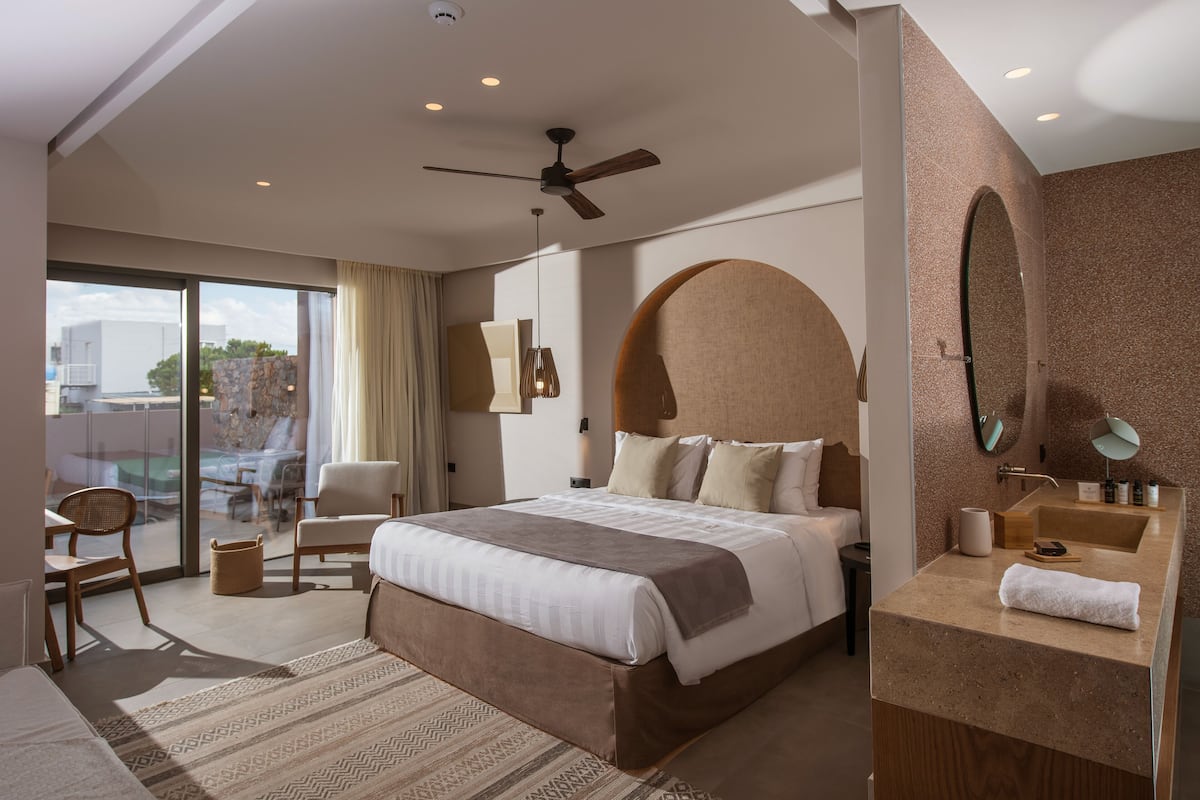 A modern hotel room with a large bed, a ceiling fan, a bathroom with a vanity, and a view of the outdoors.