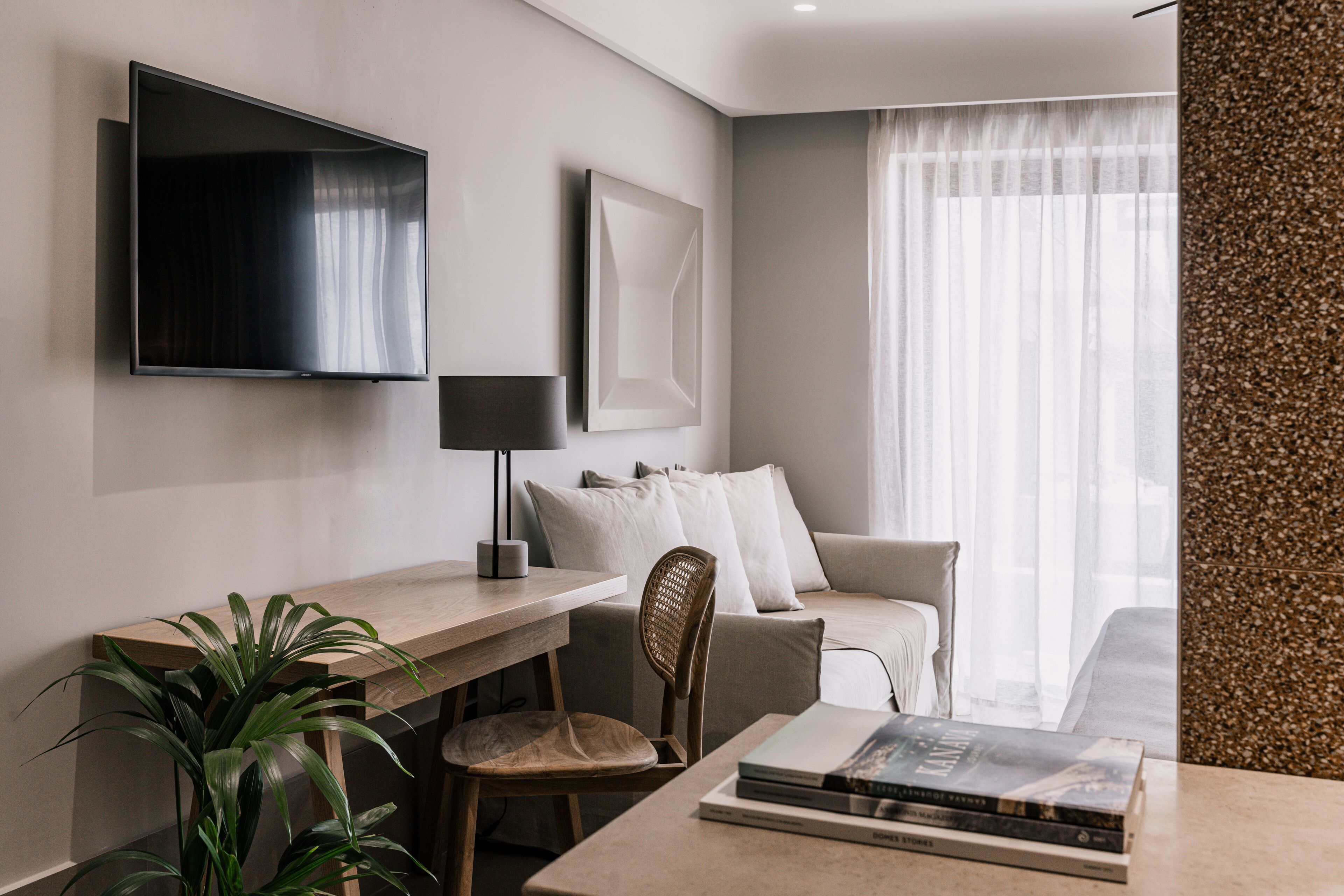 A modern hotel room with a flat-screen TV, a desk with a chair, a sofa, and a large window with sheer curtains.