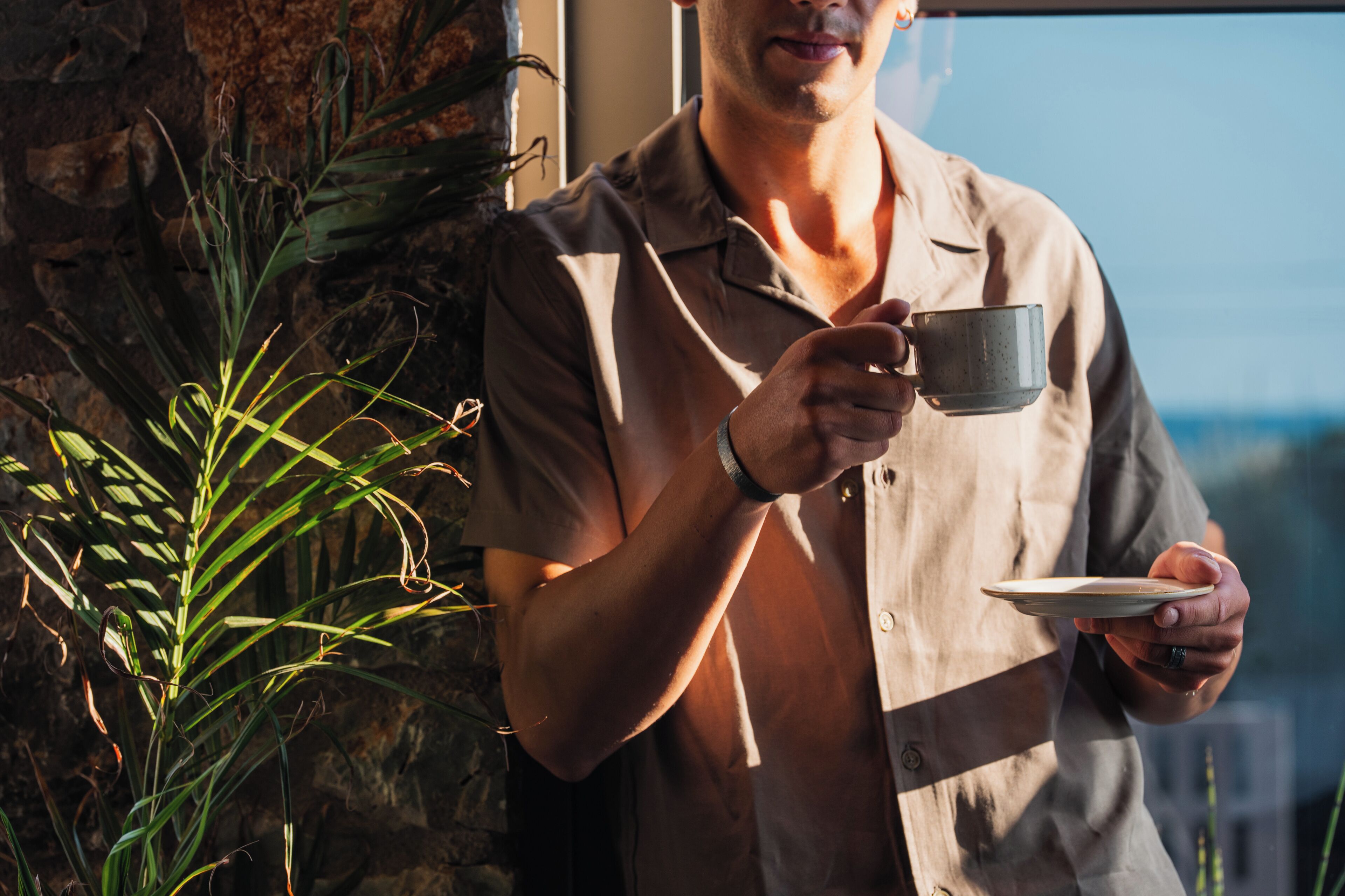 Una persona sosteniendo una taza, de pie cerca de una ventana con vista al cielo y una planta.