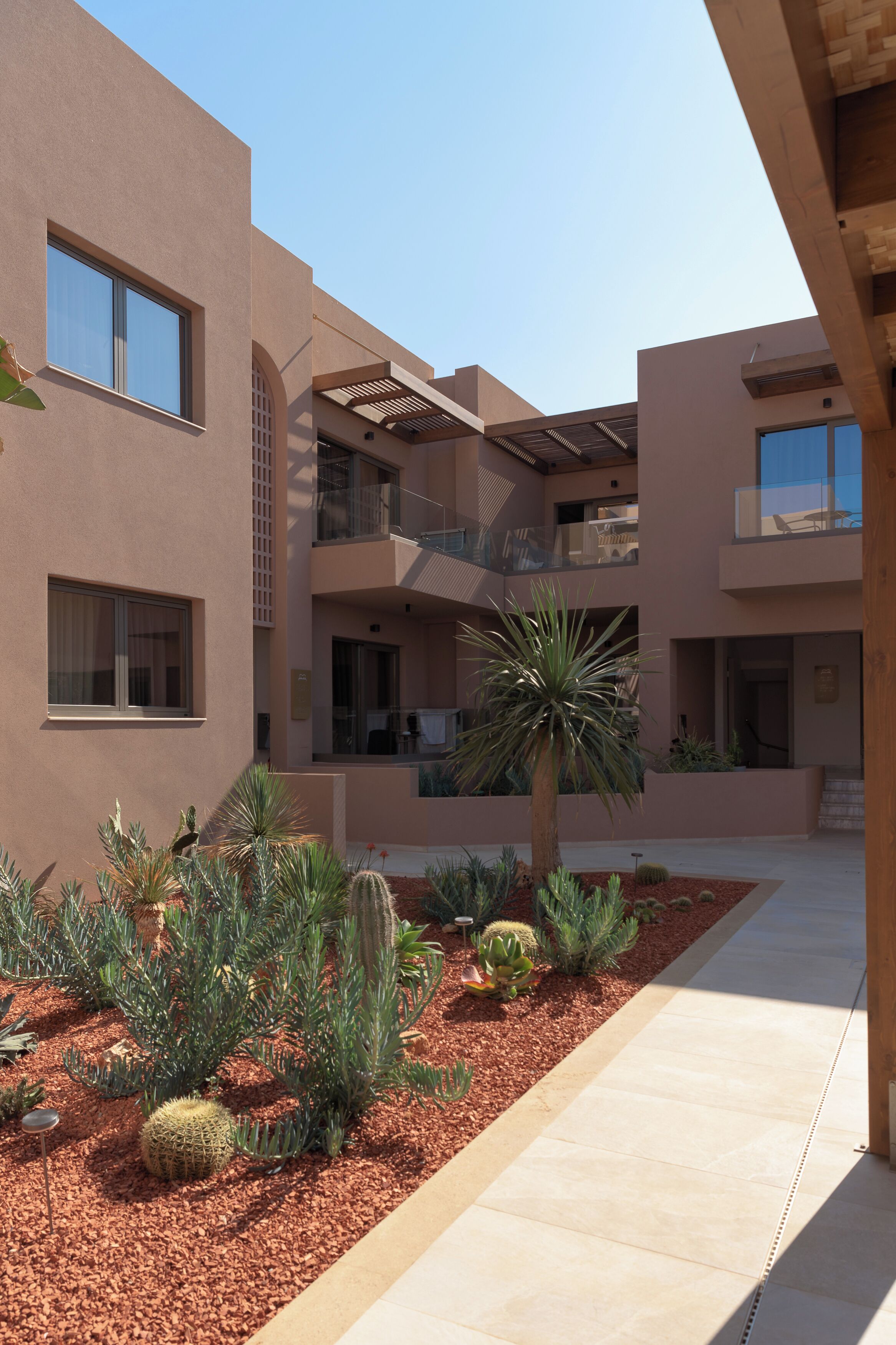 A modern building with multiple balconies, a courtyard with cacti and a palm tree, and a clear blue sky.