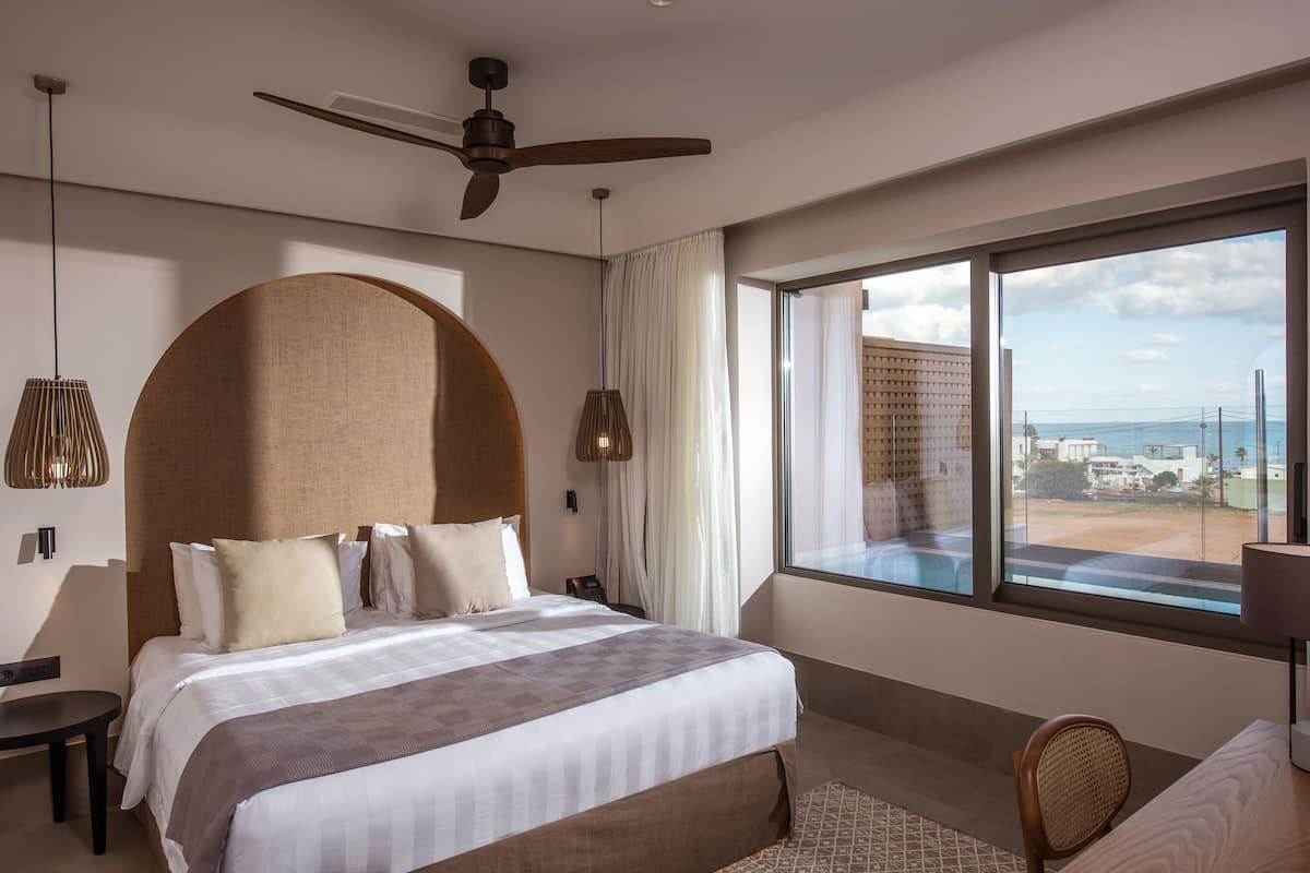 A modern hotel room with a large bed, a ceiling fan, a view of the city, and a large window.