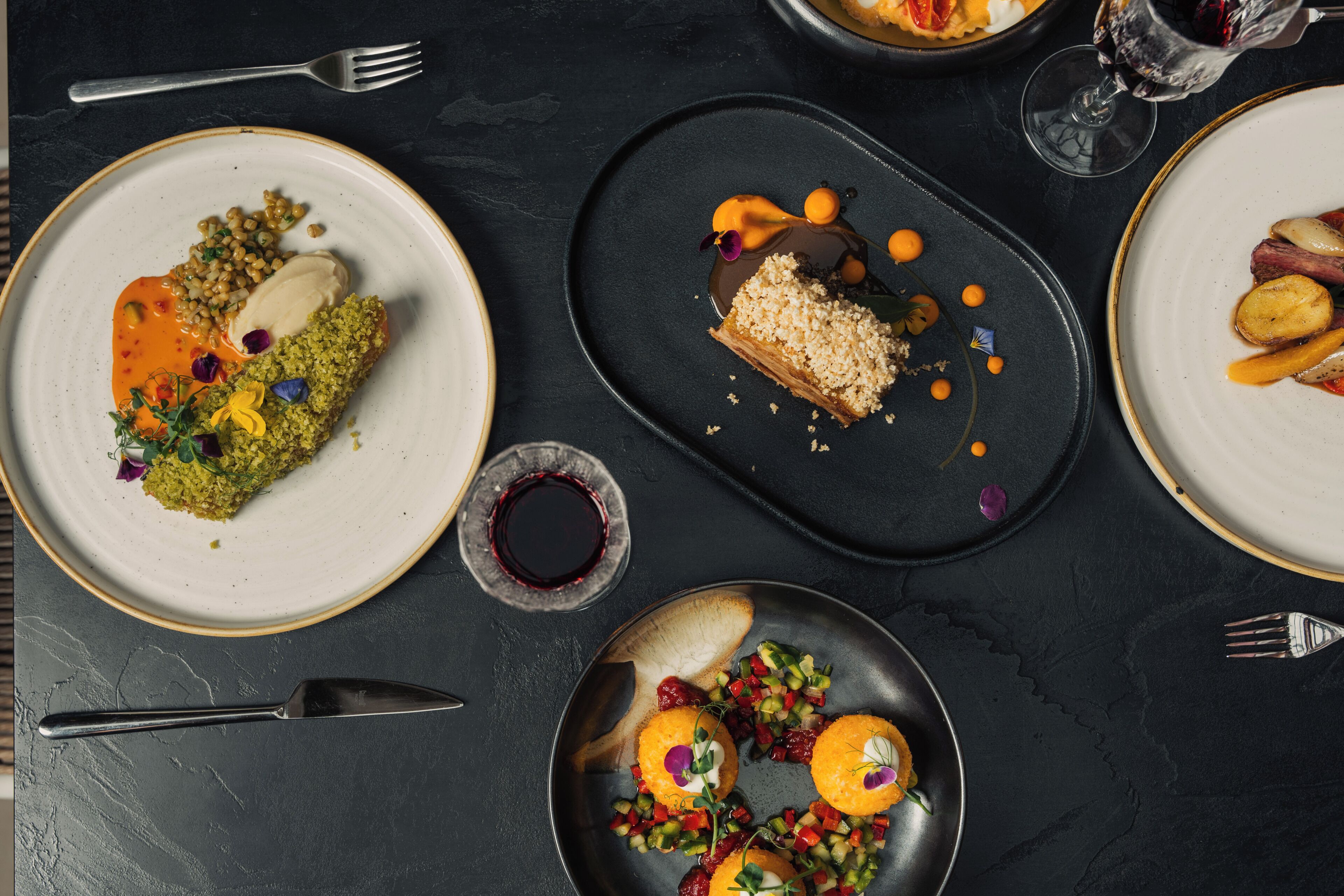 Variedad de platos en vajilla, una copa de vino y cubiertos sobre una superficie oscura.