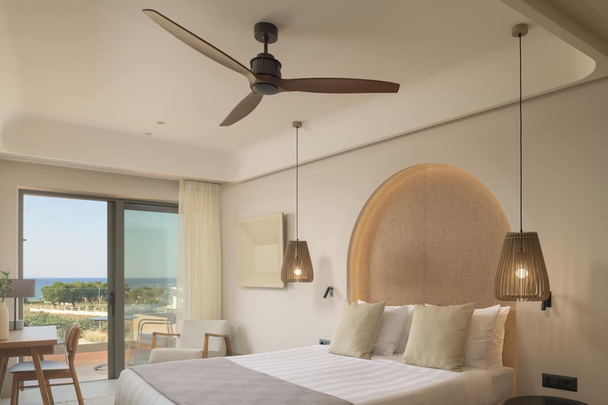 A modern hotel room with a large bed, a ceiling fan, a view of the ocean, and a dining area with a table and chairs.