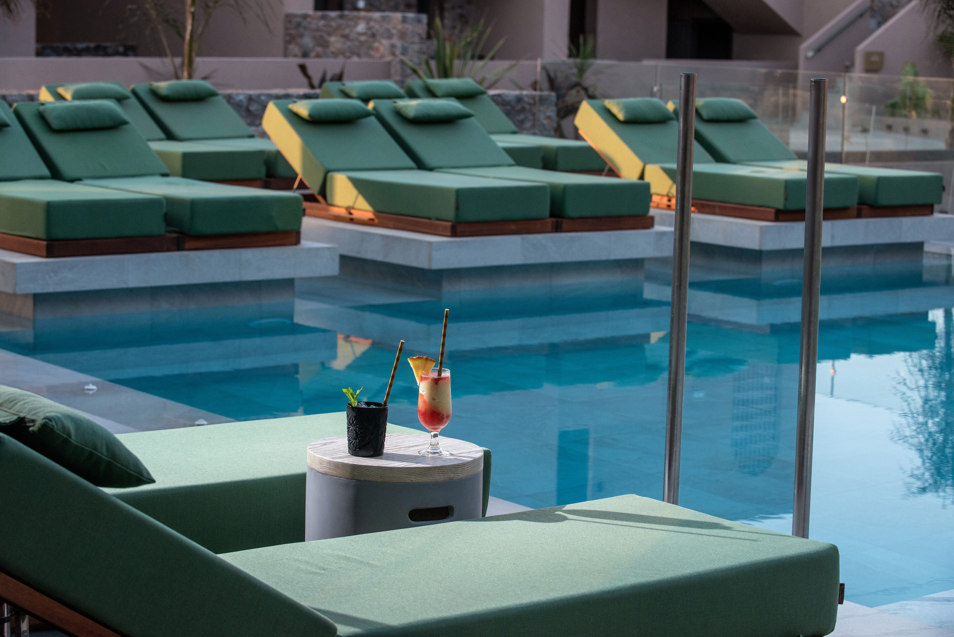 Zona de piscina con sillas reclinables verdes y una bebida sobre una mesita.