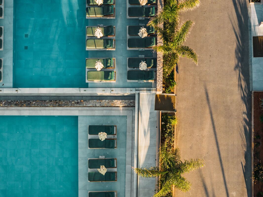 Vista aérea de un hotel con una piscina grande, múltiples zonas de descanso con sillas y sombrillas, y un camino que lleva a la piscina.