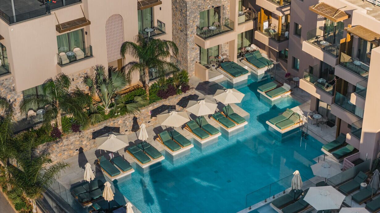 Ein Poolbereich auf dem Dach mit mehreren Liegestühlen und Sonnenschirmen, umgeben von einer Steinmauer und Bepflanzung.