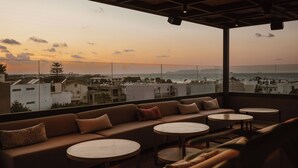 Una terraza con sofá y mesas redondas, con vistas a un paisaje urbano costero al atardecer.