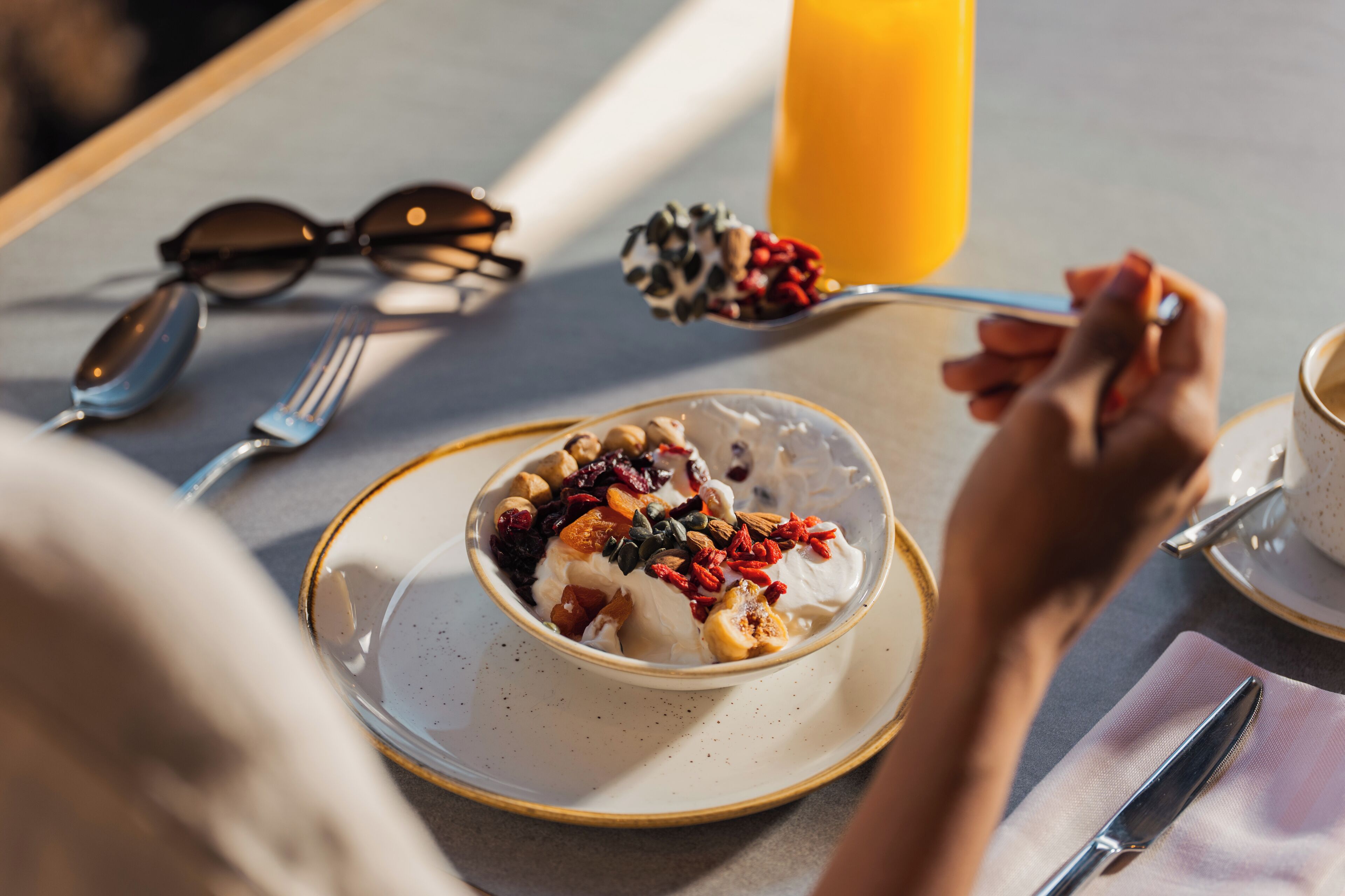 A dining table with a bowl of dessert, a spoon, a fork, a pair of sunglasses, and a glass of orange juice.