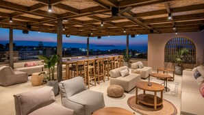 Un salón en la azotea con techo de madera, amplias ventanas y vistas del horizonte urbano al atardecer.