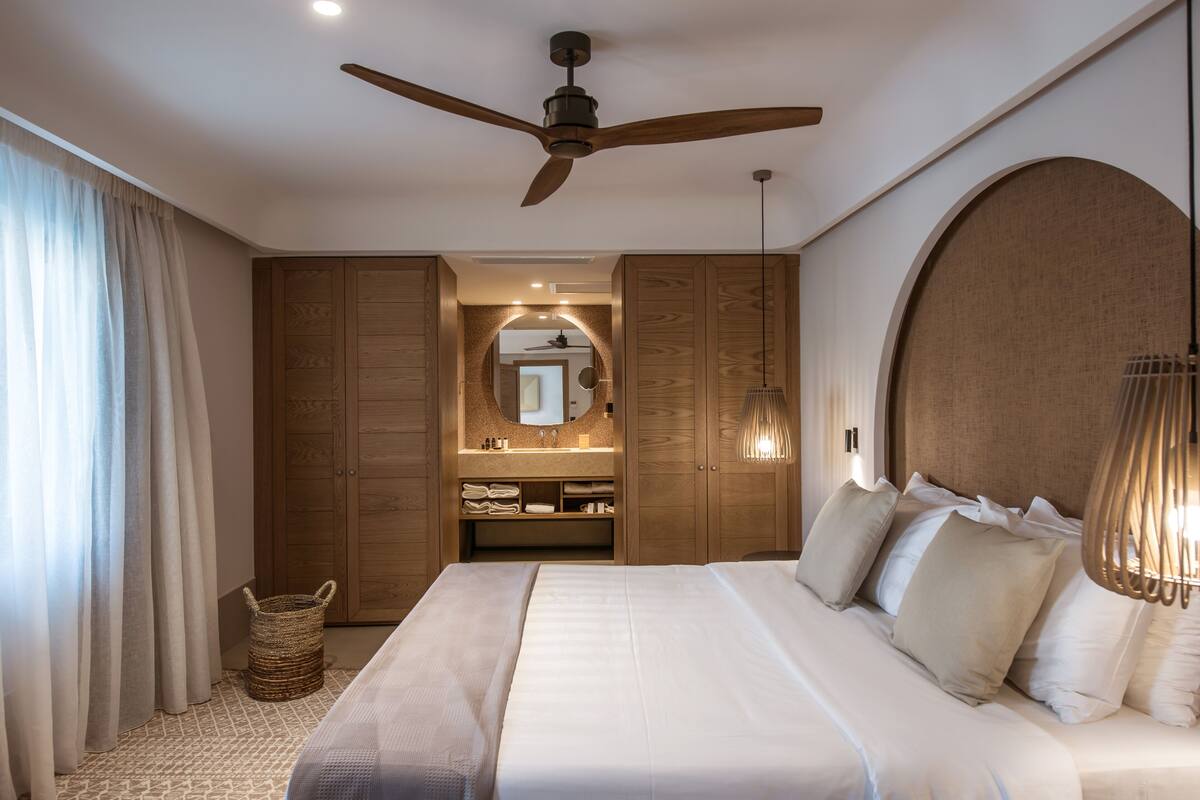A bedroom with a large bed, wooden wardrobe, a ceiling fan, a mirror, and a hanging light fixture.