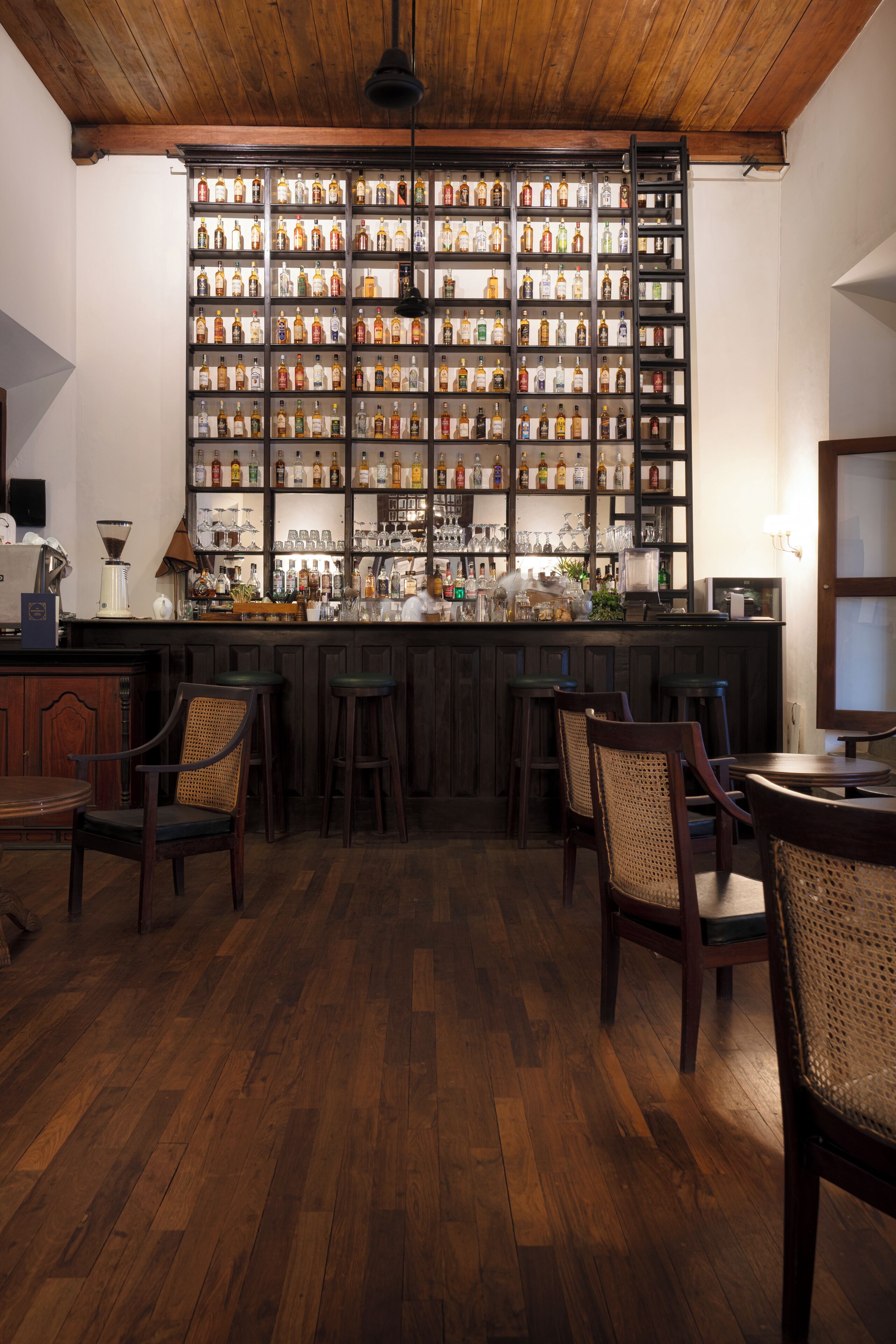 Zona de bar con suelo de madera, barra y una pared llena de botellas de licor.
