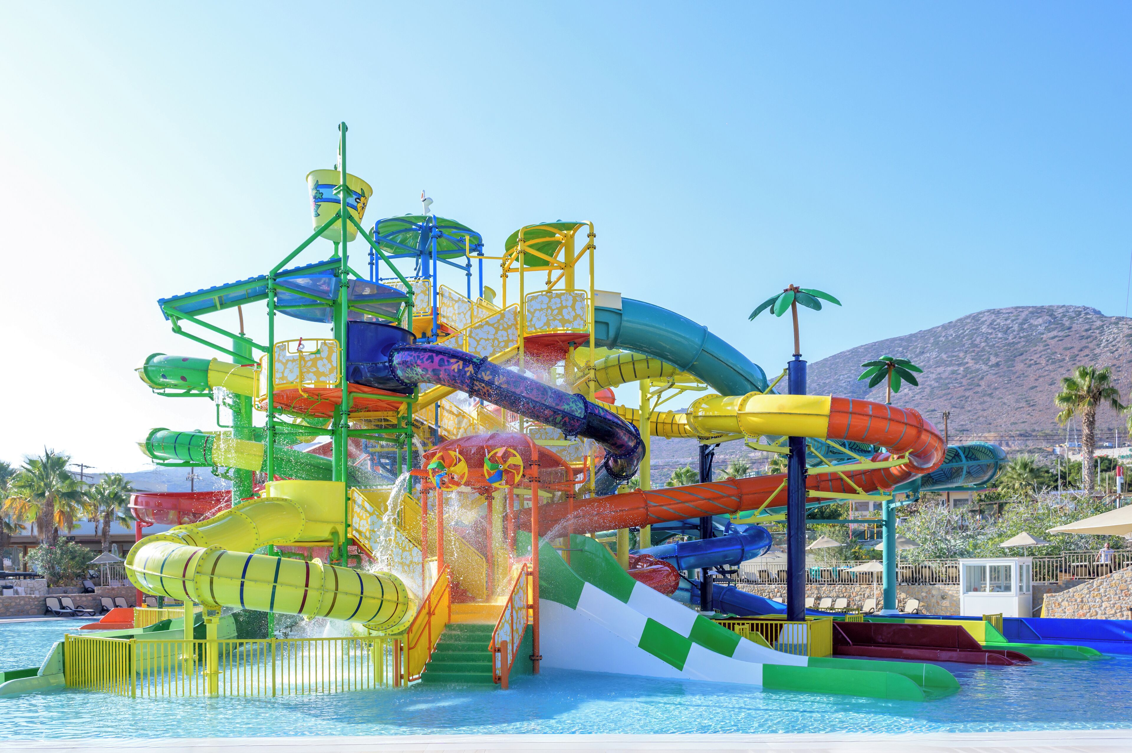 A water park with multiple slides and water features, surrounded by palm trees and a mountain in the background.