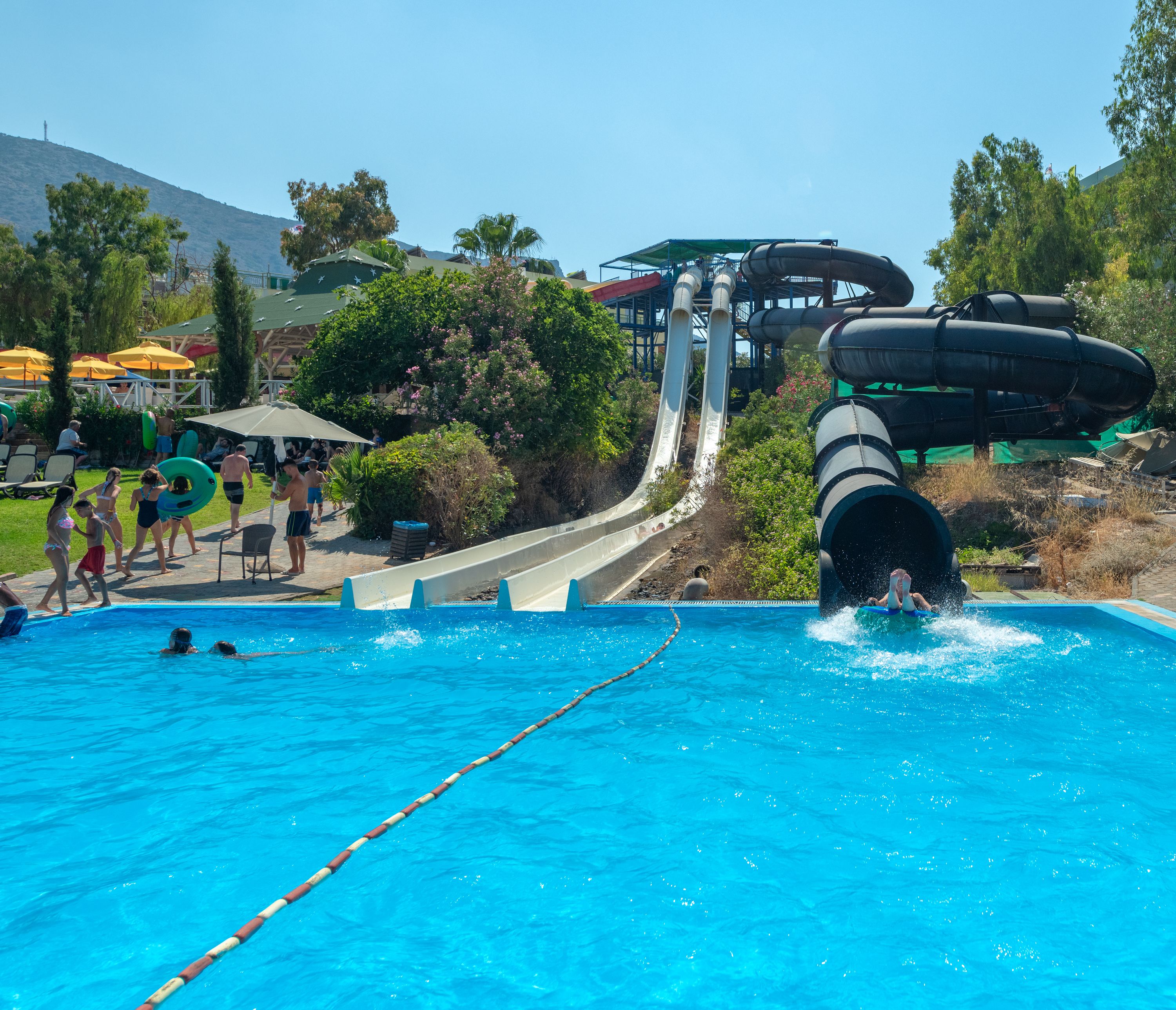 A swimming pool with a water slide, surrounded by greenery and a clear sky.