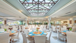 A spacious indoor dining area with round tables and wicker chairs, featuring a large glass ceiling and a view of the outdoors.