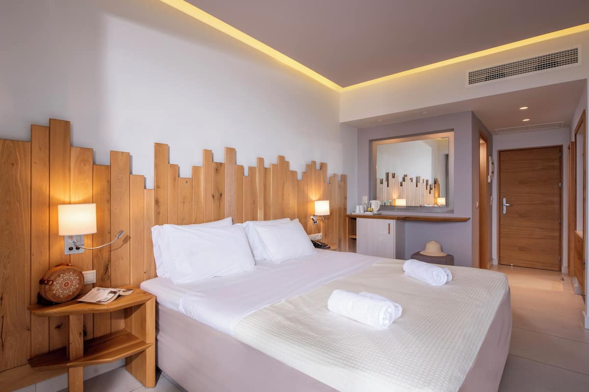 A modern hotel room with a large bed, wooden headboard, bedside lamps, a small wooden nightstand, a mirror above the bed, and a wooden door.