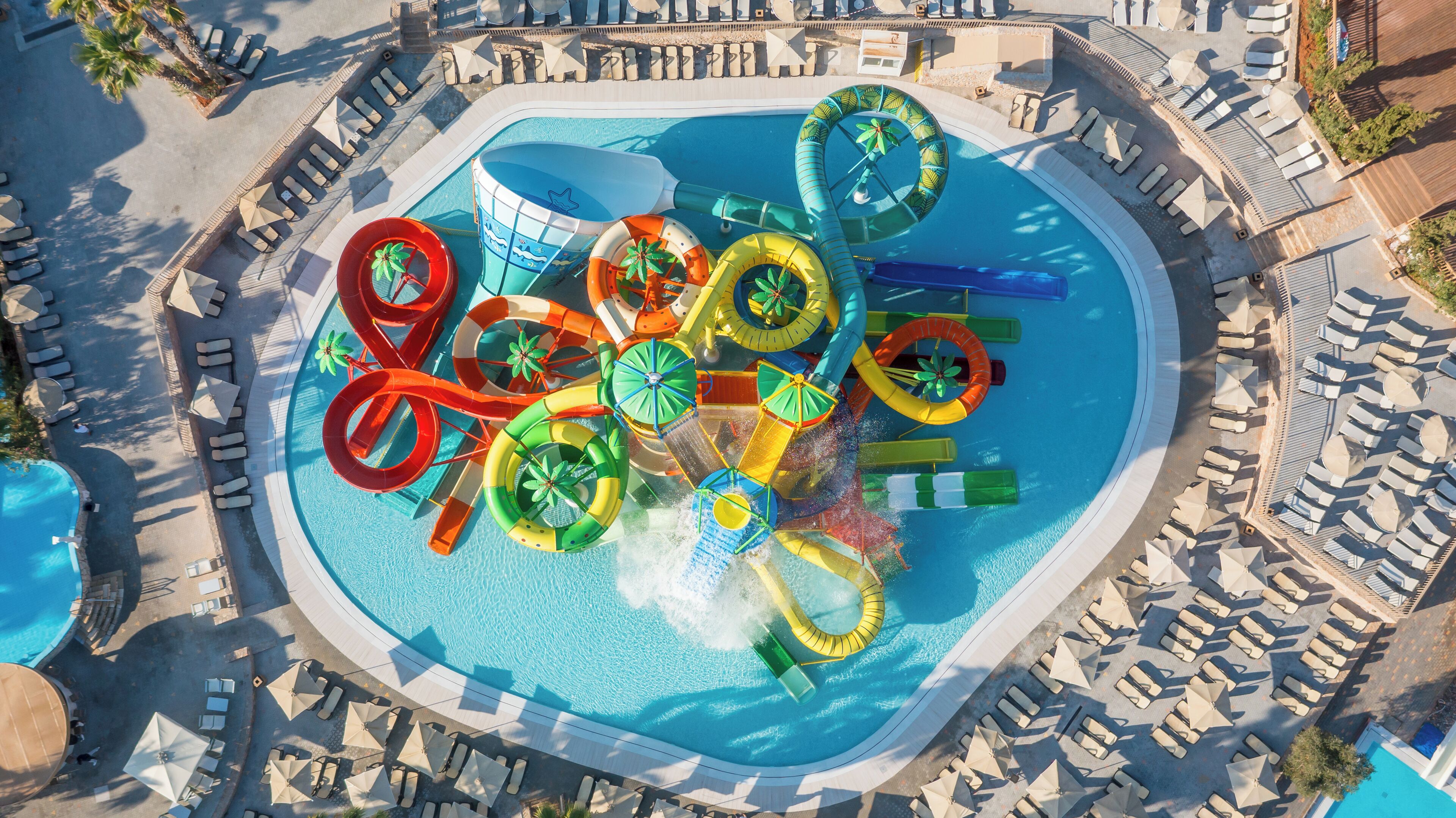Vista aérea de un parque acuático con una piscina grande, varias toboganes de agua y tumbonas dispuestas alrededor de la zona de la piscina.