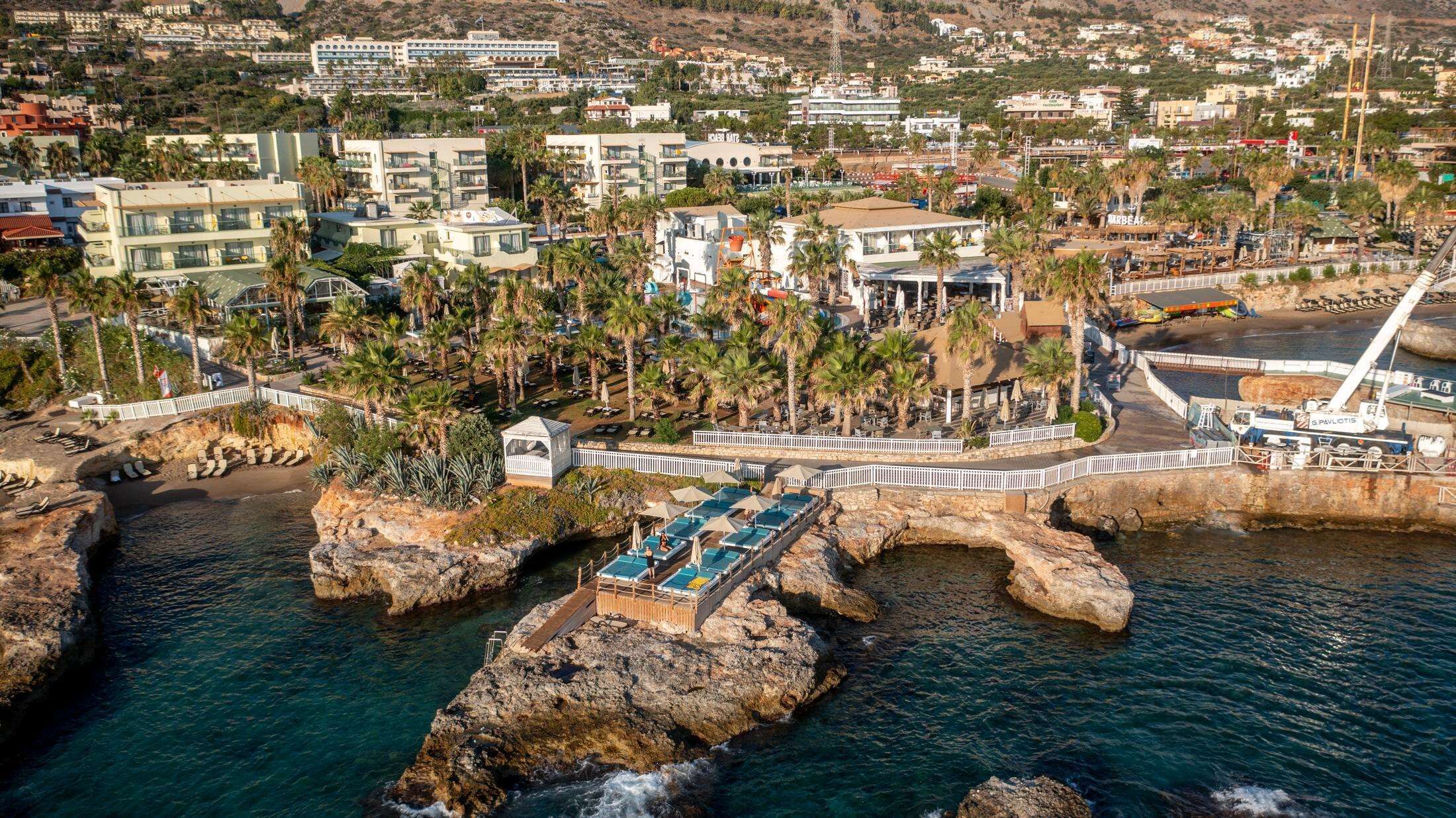 A coastal hotel with a rocky shoreline, a pool area, and a view of the city in the background.