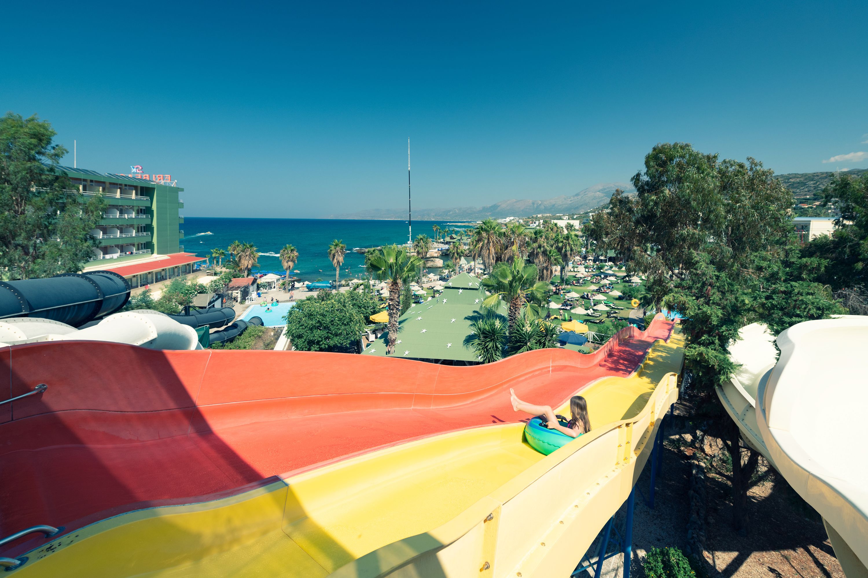 A water park with a large red and yellow slide, palm trees, and a view of the ocean.