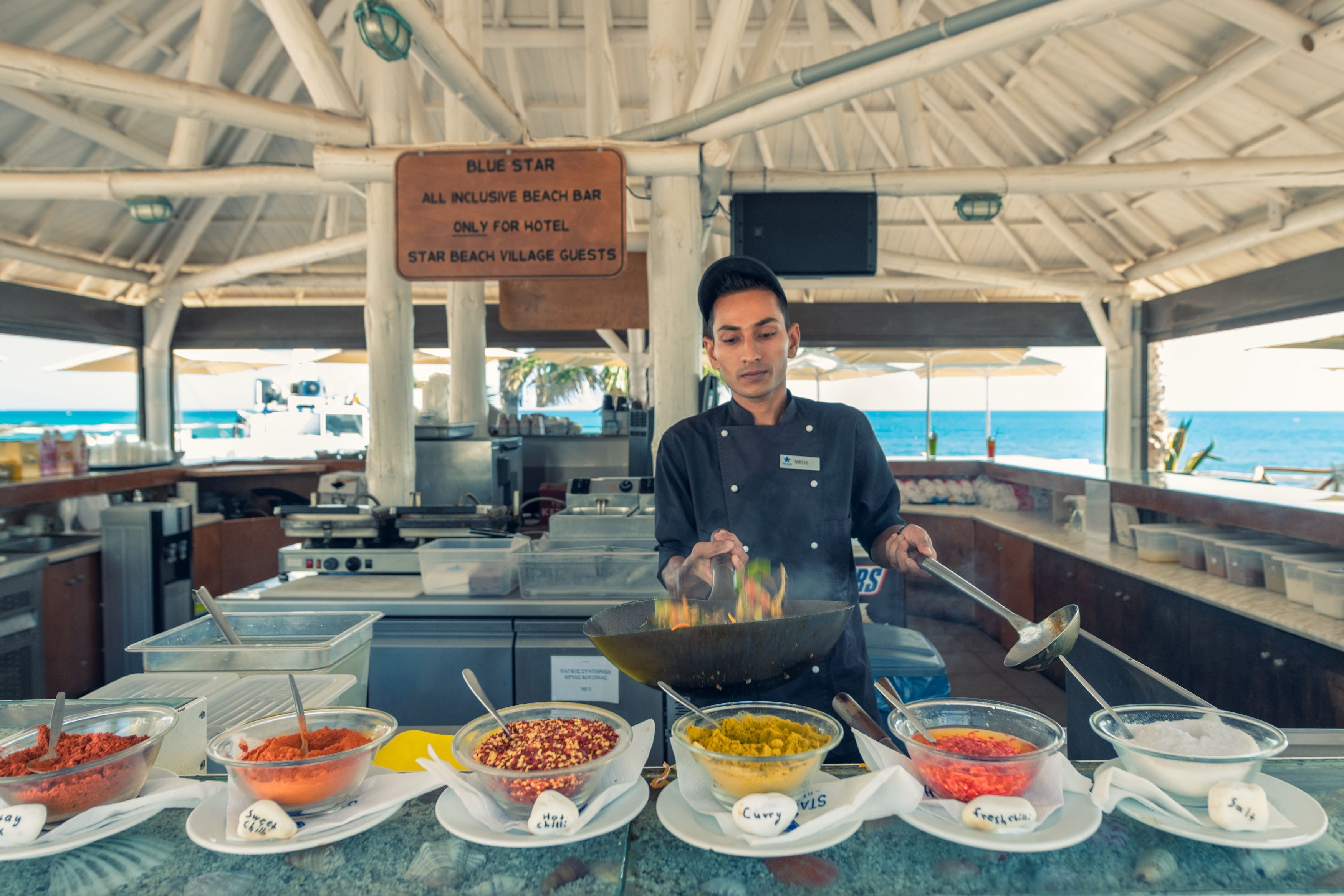 Een kok aan het koken in een overdekte ruimte met uitzicht op het strand, diverse ingrediënten op het aanrecht, en een bord dat aangeeft dat de locatie voor hotelgasten is.